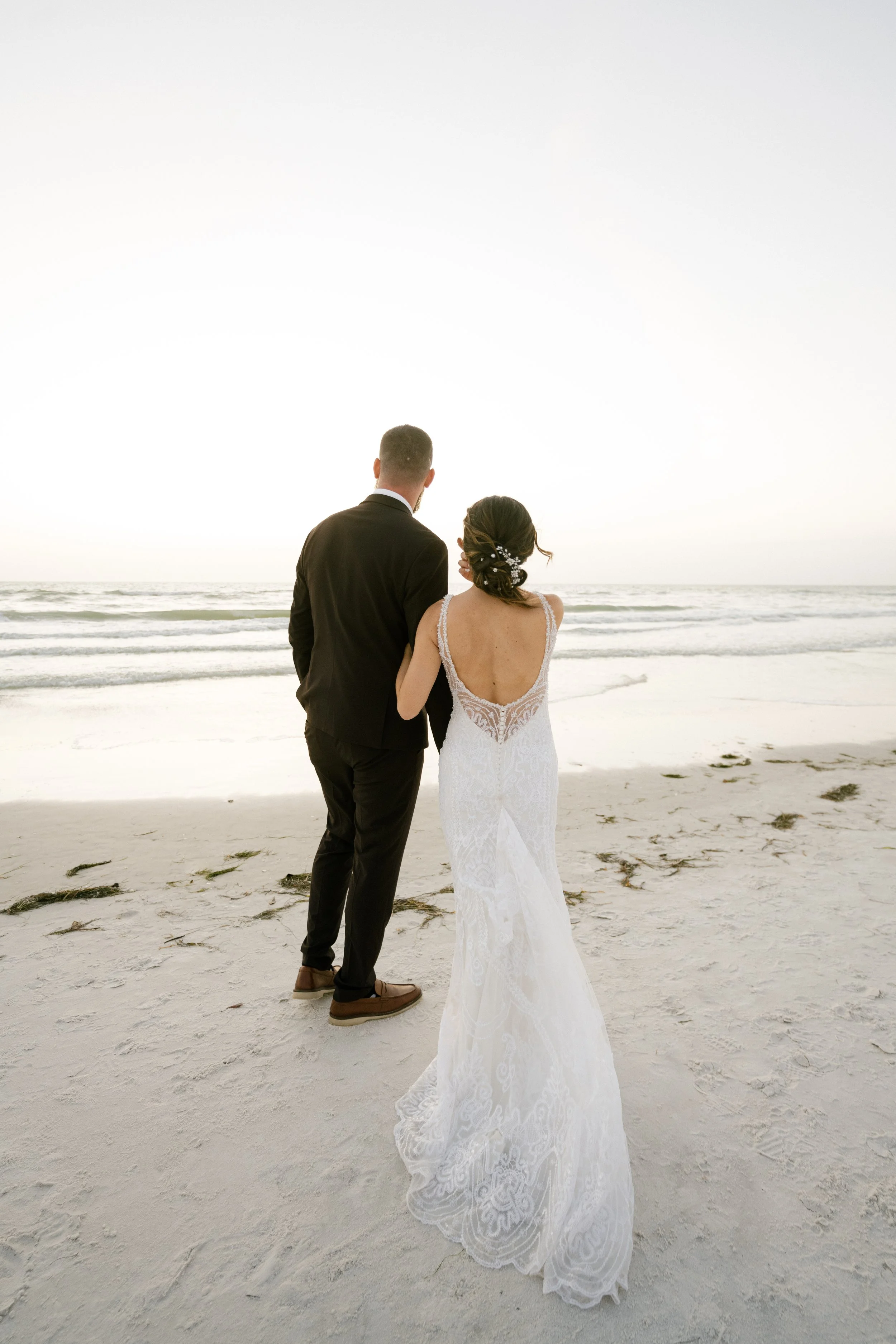 A bride and groom walk on a beach at sunset, holding hands and enjoying a romantic moment.