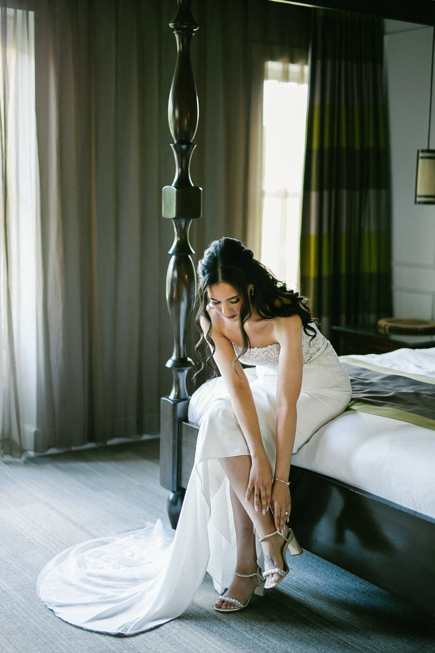 A bride in a white wedding dress sitting on a bed, adjusting her shoes.