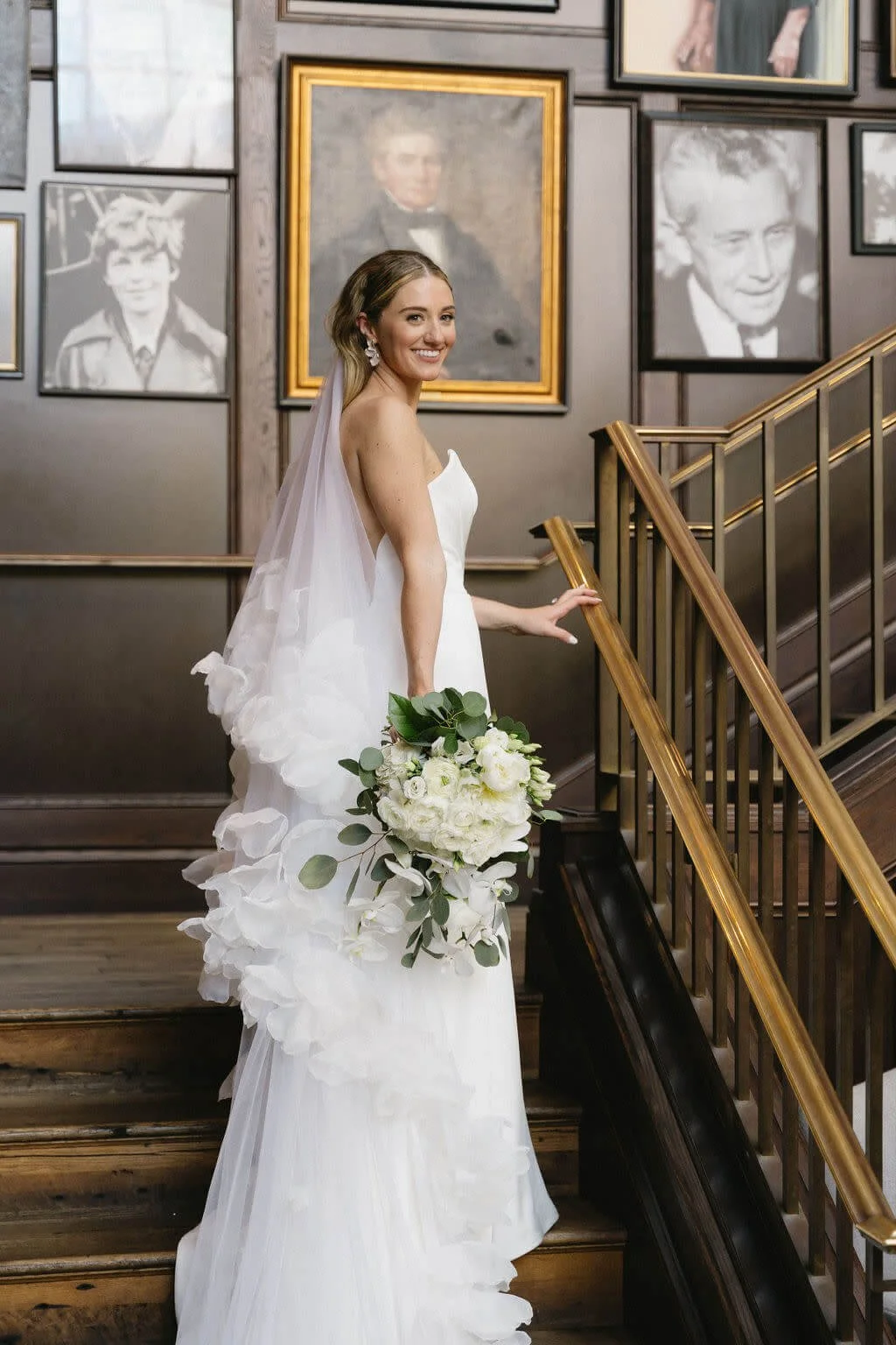 Bride in white wedding gown with bouquet on wooden staircase surrounded by framed black-and-white portraits on the wall.