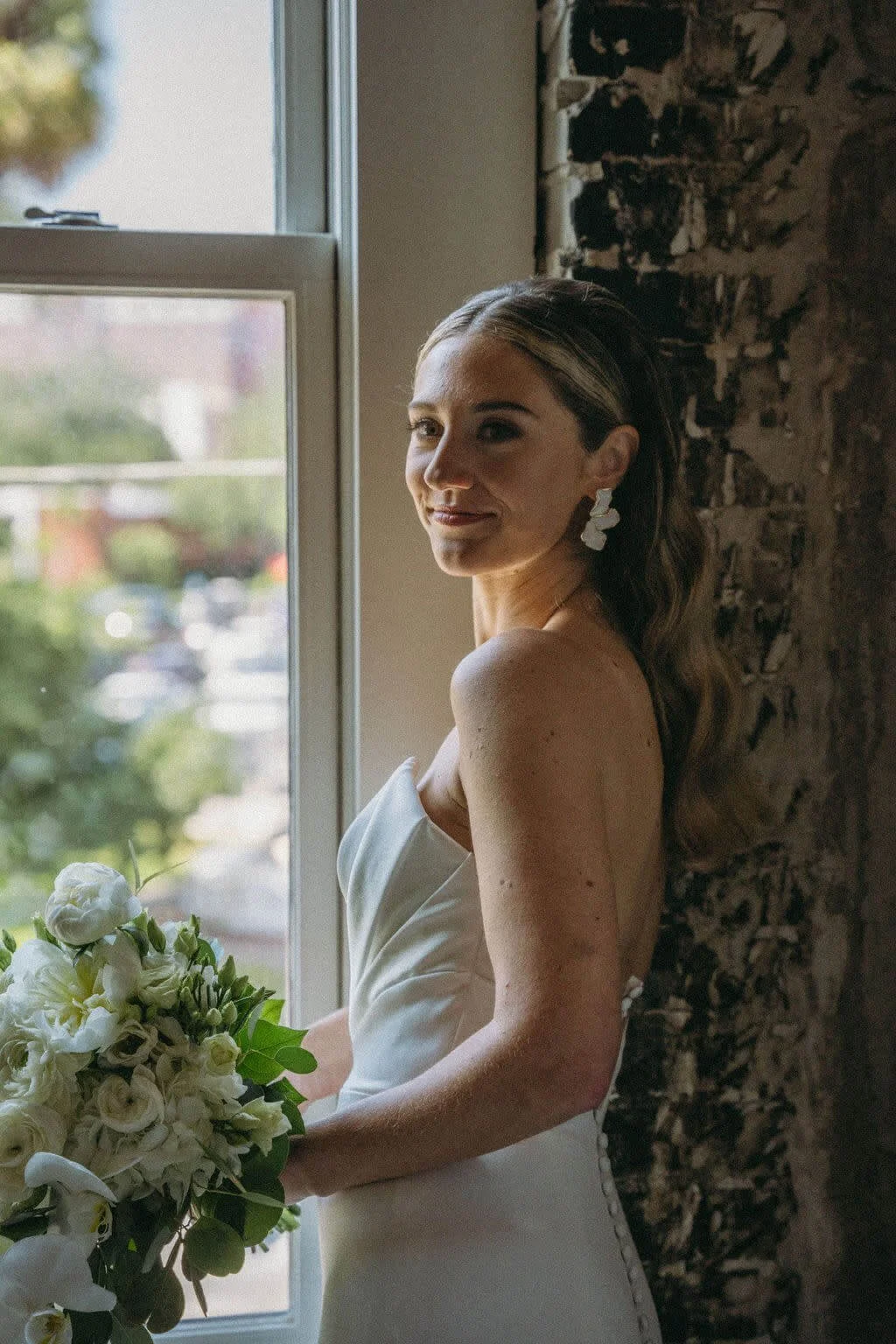 A bride standing near a window holding a bouquet of white flowers, wearing a strapless white wedding dress, with a textured brick wall in the background.