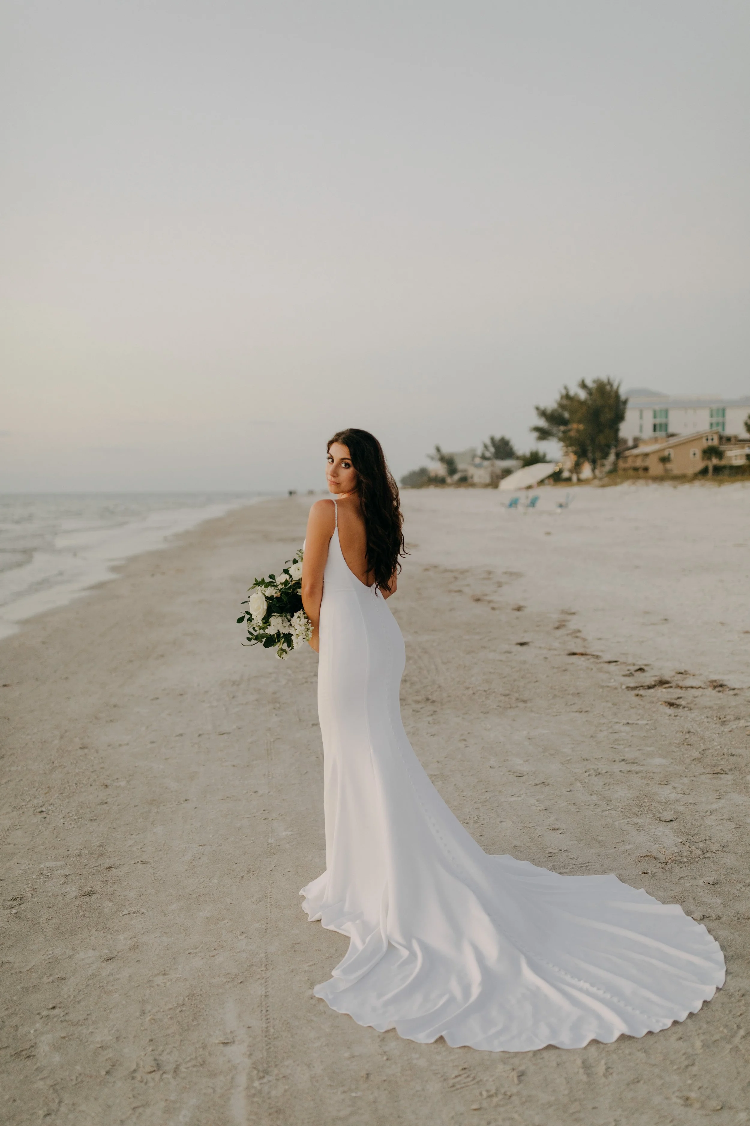 Bride standing on the beach holding a bouquet of flowers, with houses and trees in the background.