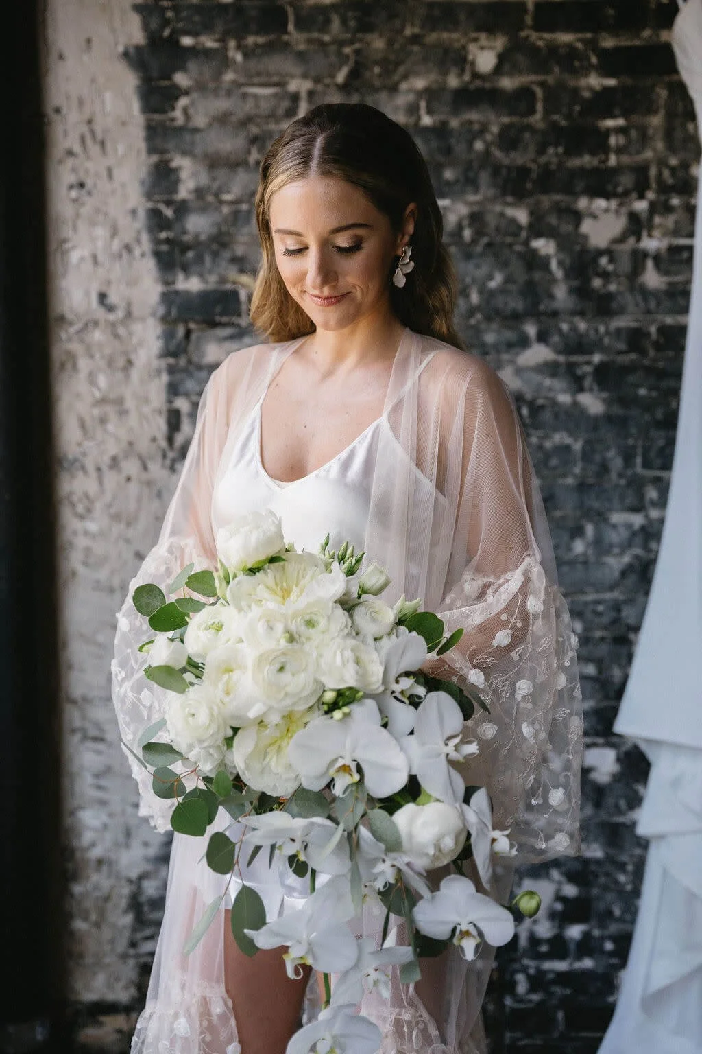 A bride holding a large bouquet of white flowers, standing against a rustic brick wall, wearing a white slip dress covered by a sheer, embroidered lace robe, with earrings and makeup.