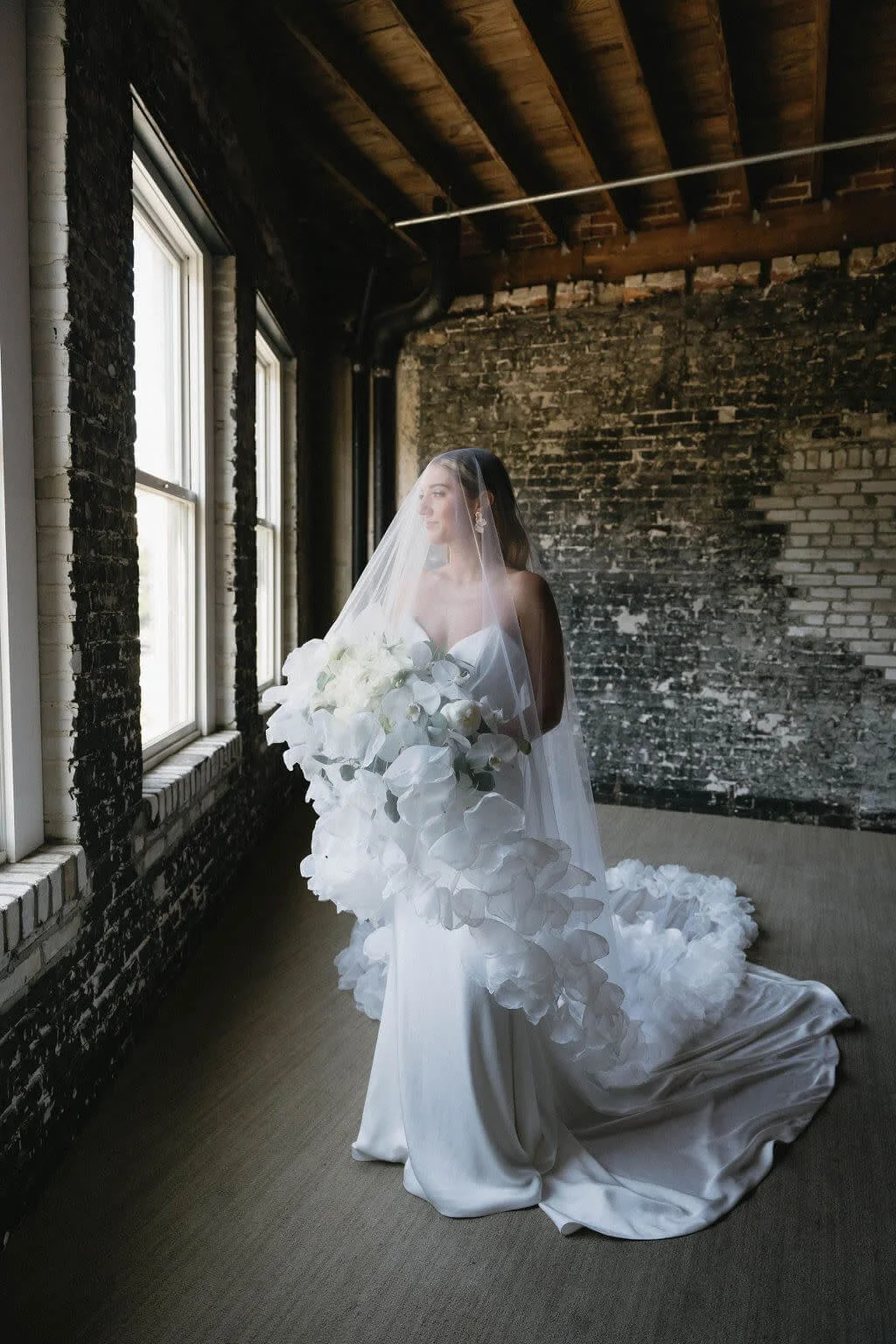 A bride in a white wedding gown and veil holding a large bouquet of white flowers, standing in a rustic room with brick walls and wooden ceiling beams, looking out the window.
