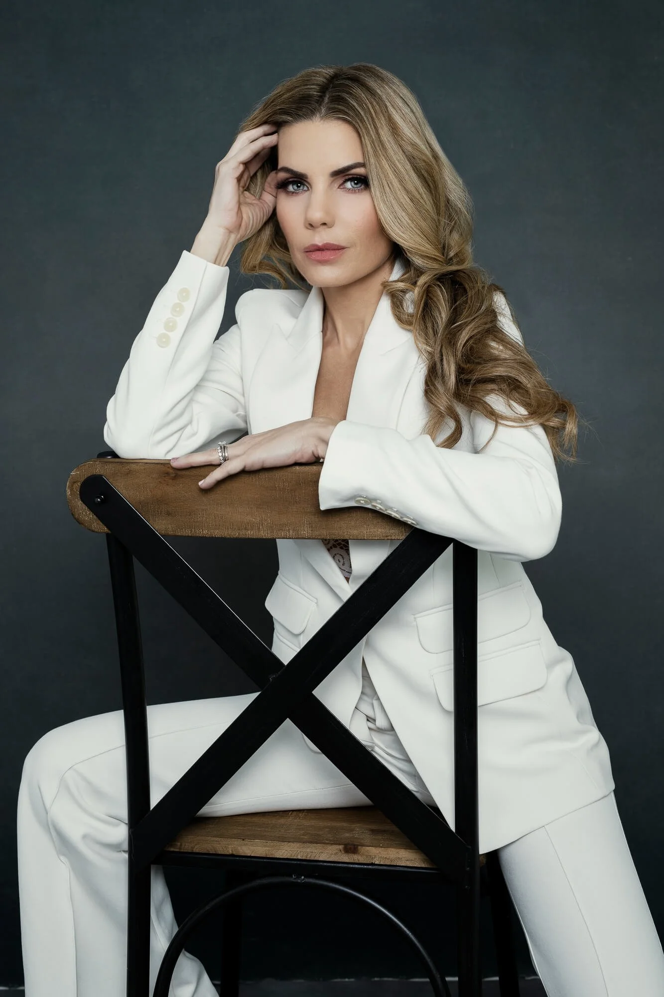 A woman with long, wavy blonde hair wearing a white suit, sitting backward on a wooden and metal chair, resting her arms on the chair's backrest, looking confidently at the camera against a plain gray background.