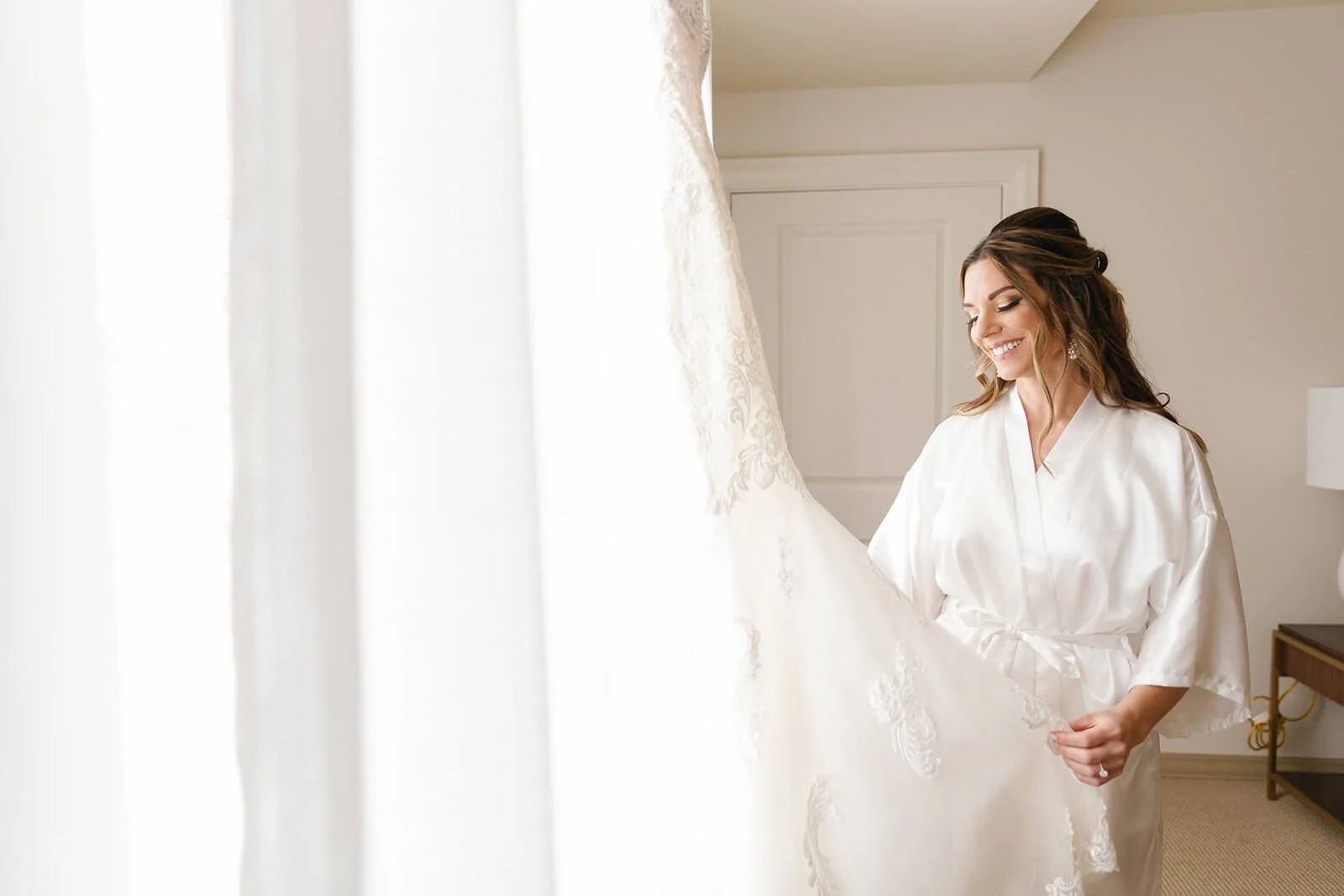 A woman in a white satin robe smiling while holding her wedding dress in a bright room.