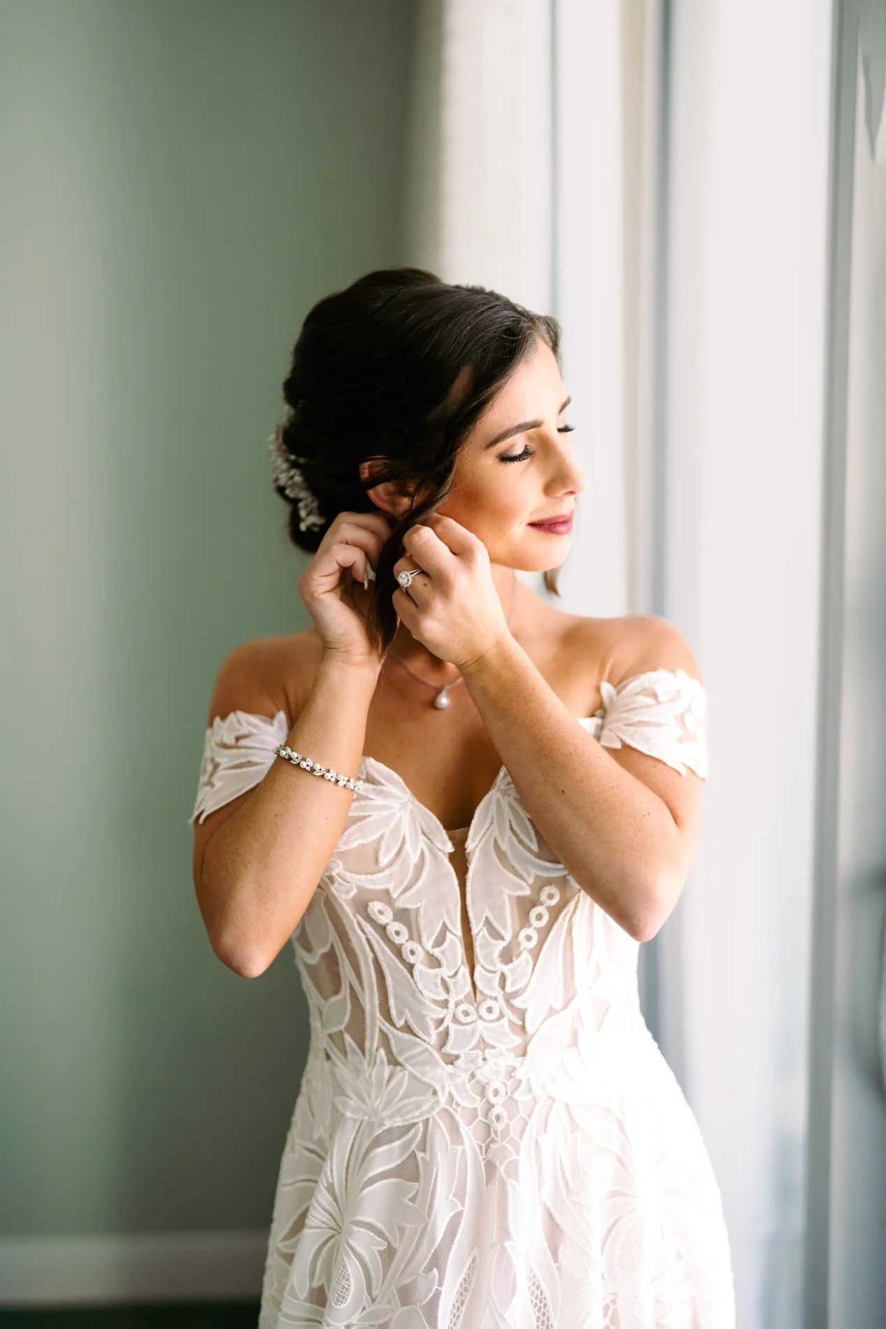 A bride with dark hair styled in an updo, wearing an off-the-shoulder white lace wedding dress, placing earrings while standing near a window with soft natural light.