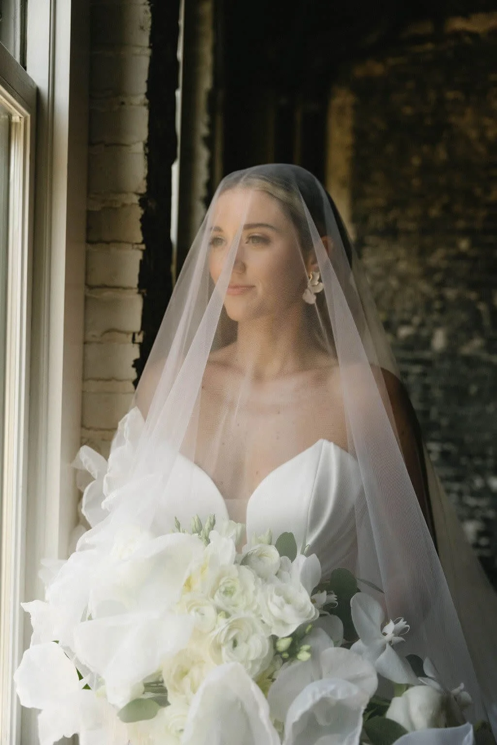 A bride with a veil holding a bouquet of white flowers, standing by a window.