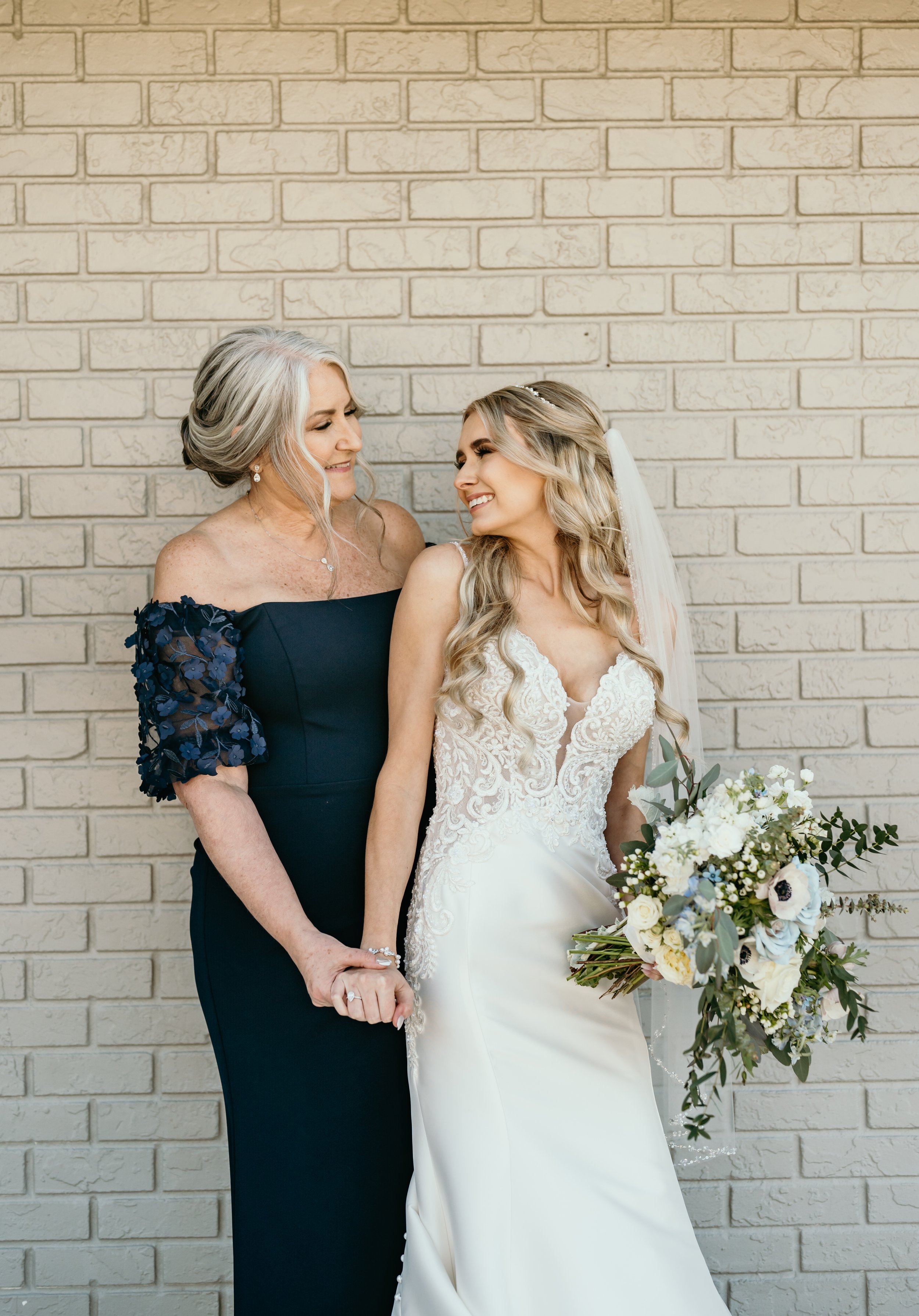 A wedding bride and her mother holding hands and smiling in front of a beige brick wall. The bride is in a white lace wedding dress with a bouquet of white and green flowers. The mother is in a navy blue dress with lace sleeves.