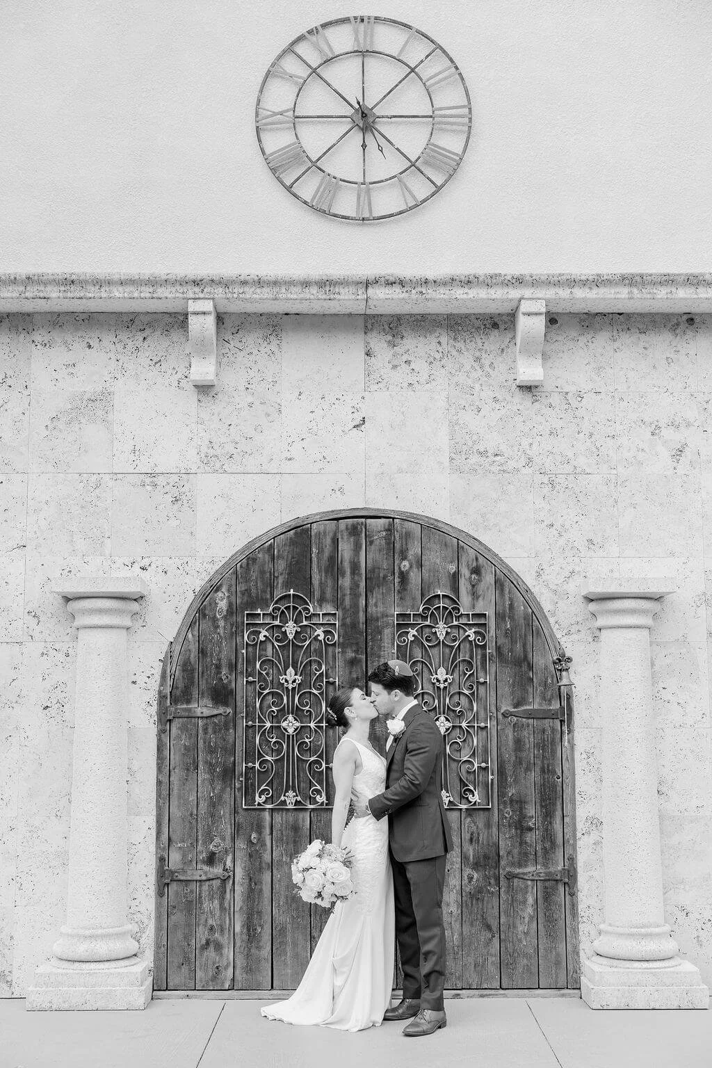 Black and white photo of a newlywed couple sharing a kiss in front of a wooden door with ornate metalwork, with a large clock mounted on the wall above them.