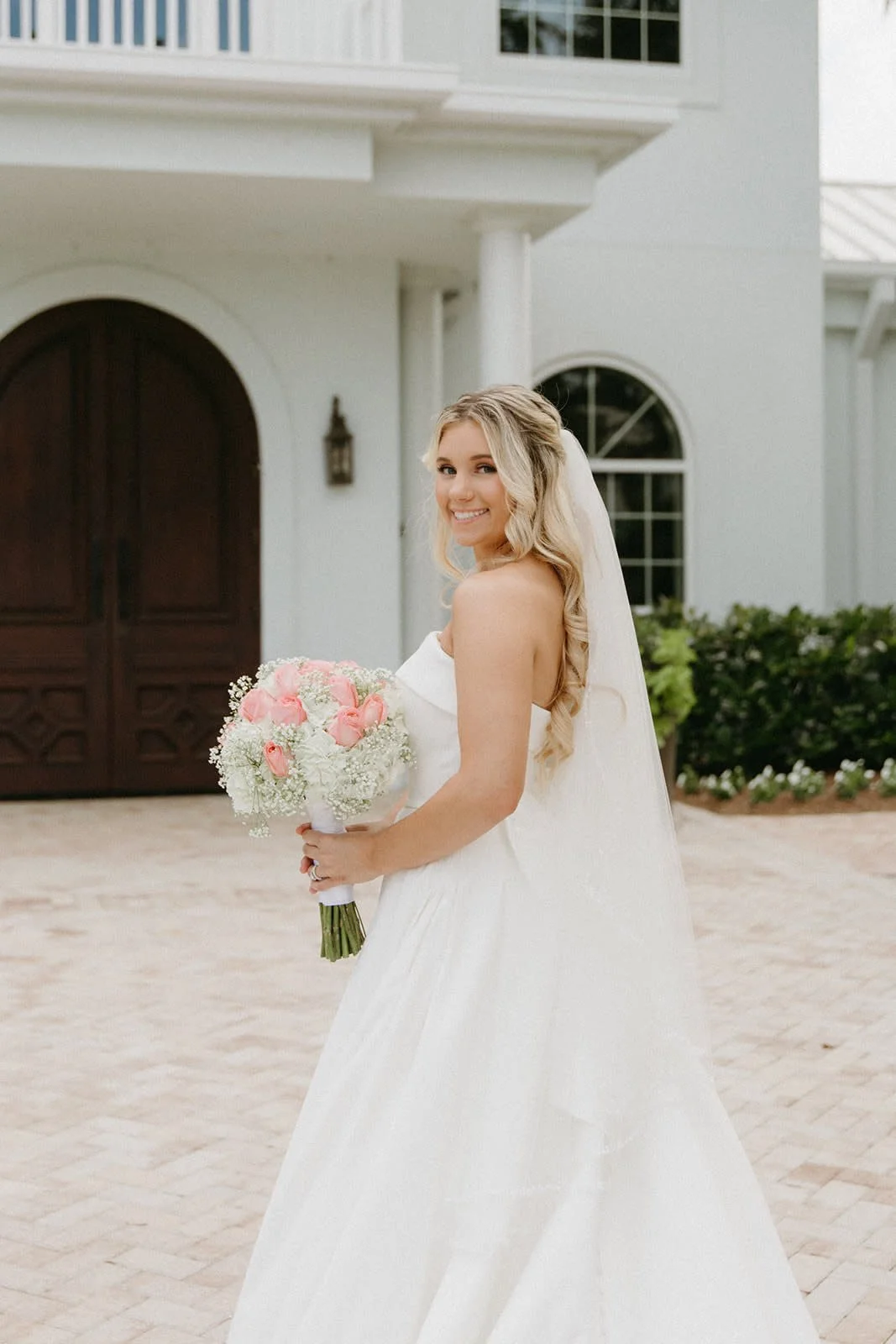 A smiling bride with blond hair, wearing a white wedding dress and veil, holding a bouquet of pink and white flowers, standing outside a white building with dark wooden doors and arched windows.
