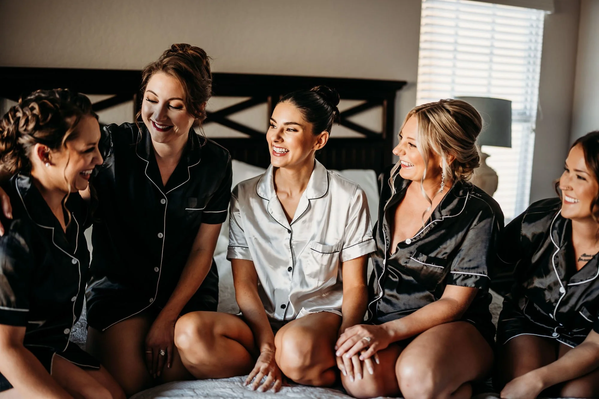 Bridal party sitting on a bed, wearing matching satin pajamas, smiling and laughing together in a bedroom.