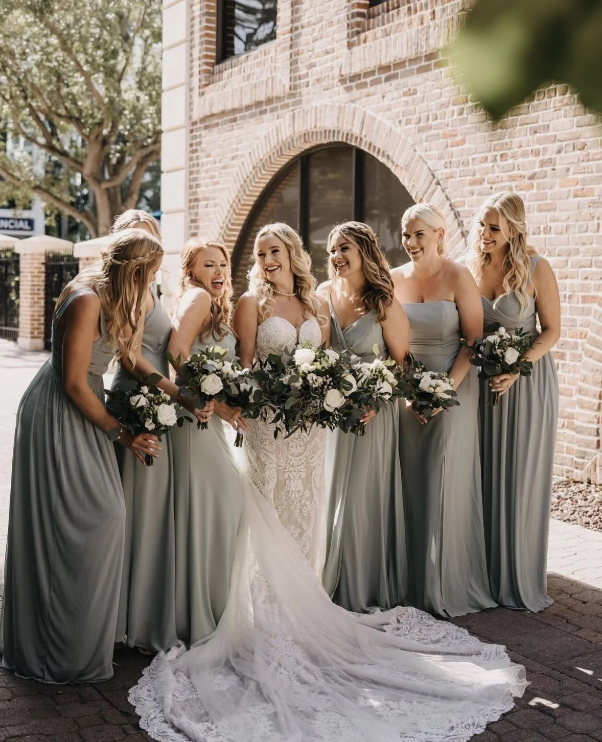 A bride and six bridesmaids standing outside near a brick building, all smiling and holding bouquets of white flowers and greenery.