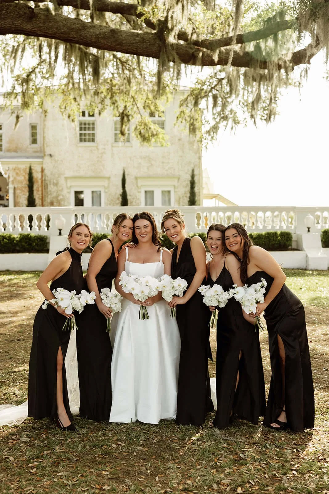 A bride and five bridesmaids in black dresses holding white floral bouquets, standing outdoors in front of a white stone building and large tree with moss, smiling.