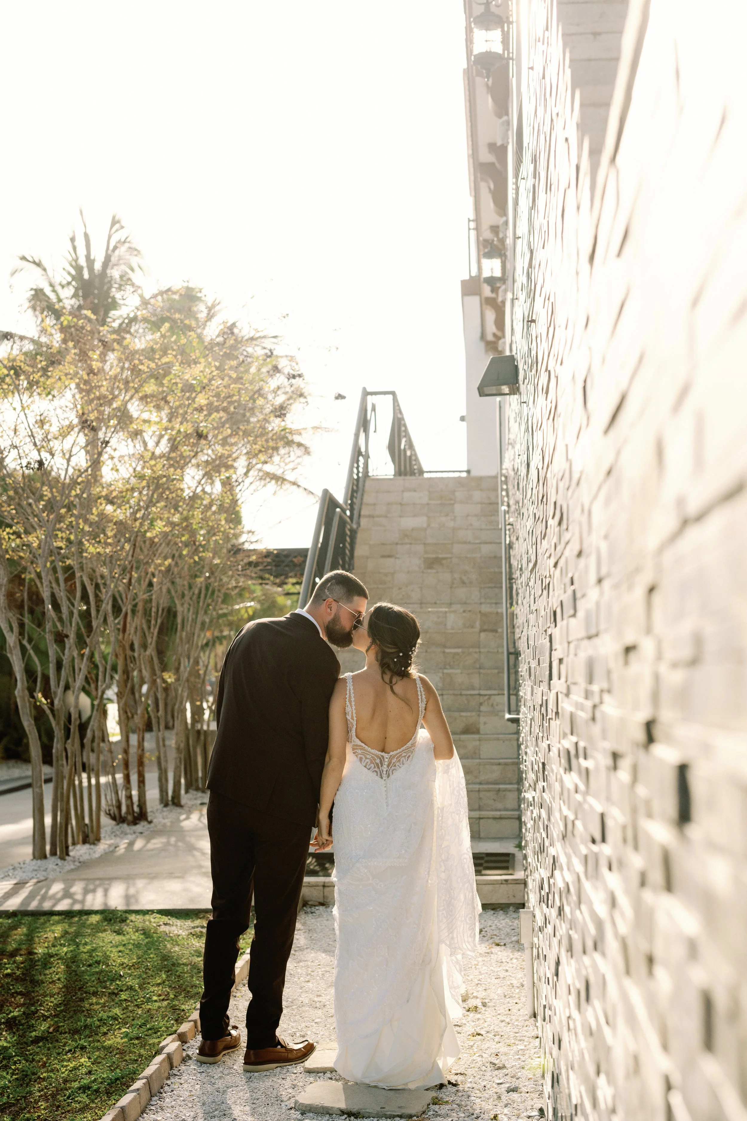 A bride and groom holding hands, standing close and touching foreheads, outdoors near a staircase and brick wall, during daylight.