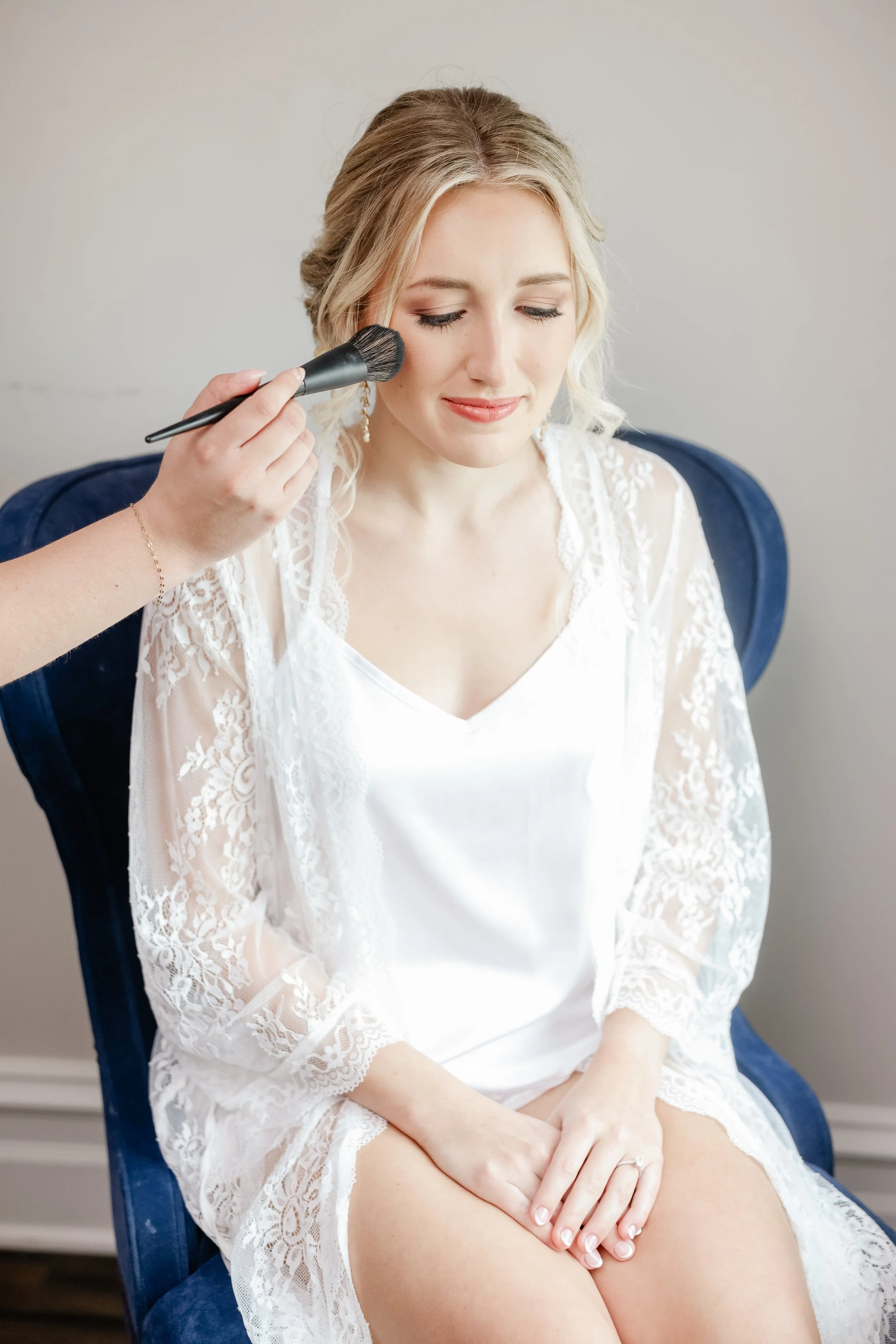 A woman with blonde hair getting makeup applied to her face with a brush, sitting on a blue chair, wearing a white lace robe and a white slip.