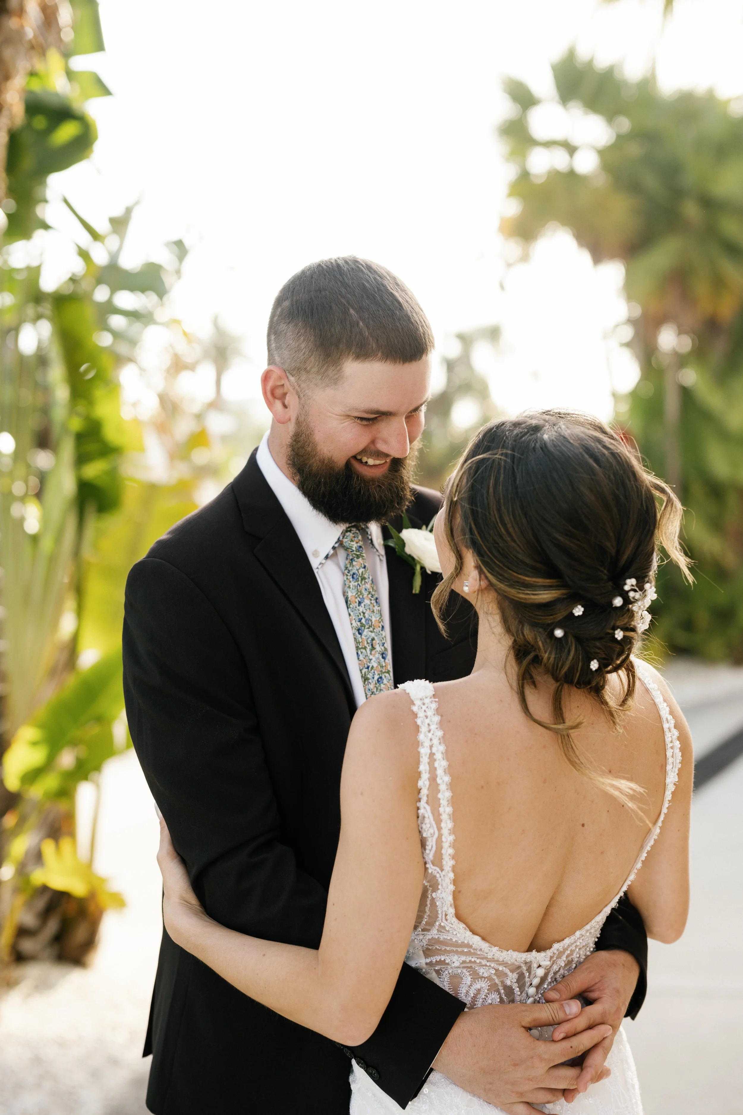 A newlywed couple is smiling and embracing outdoors, with  trees in the background, during their wedding celebration.