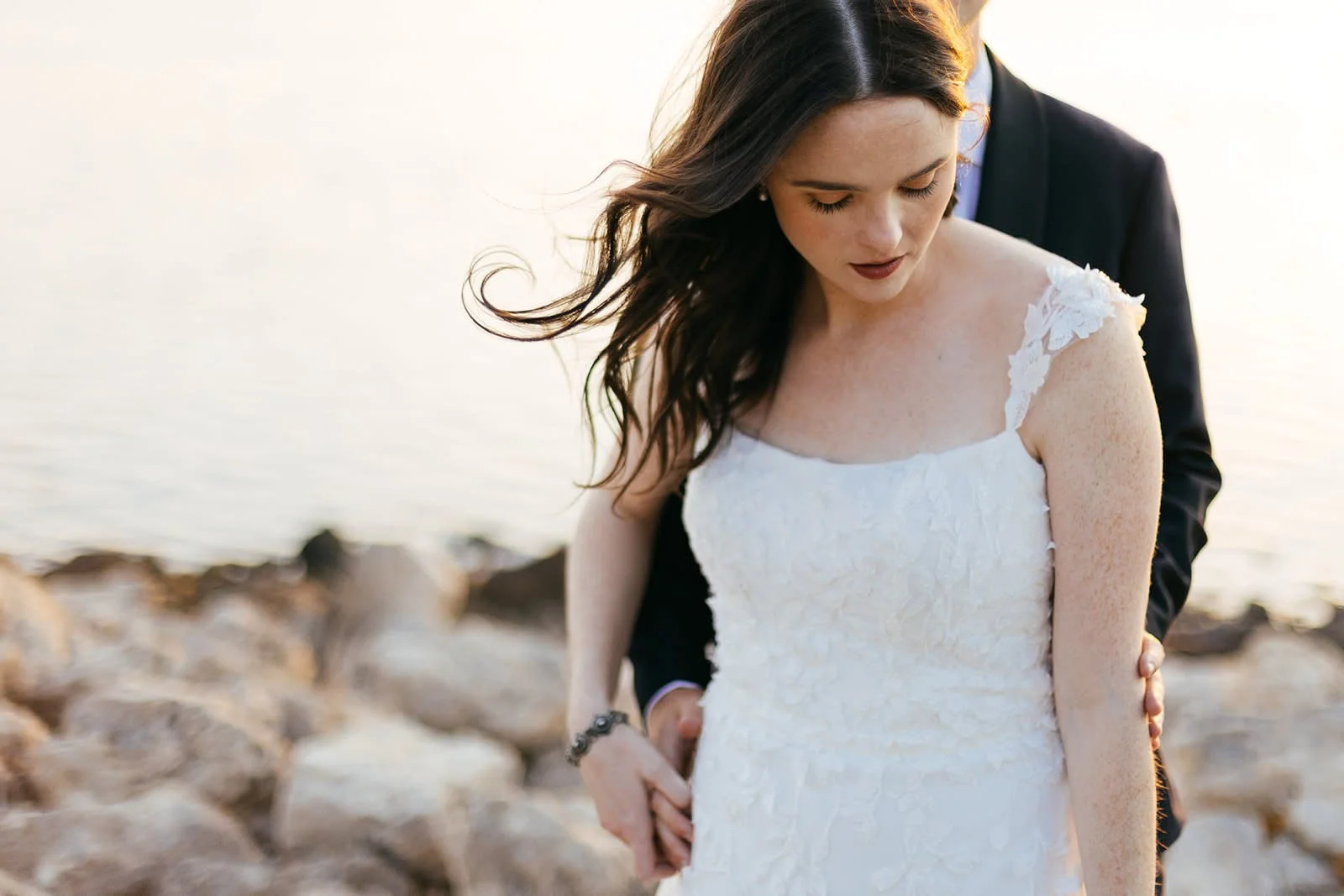 Bride in white lace wedding dress embraced by groom on rocky beach