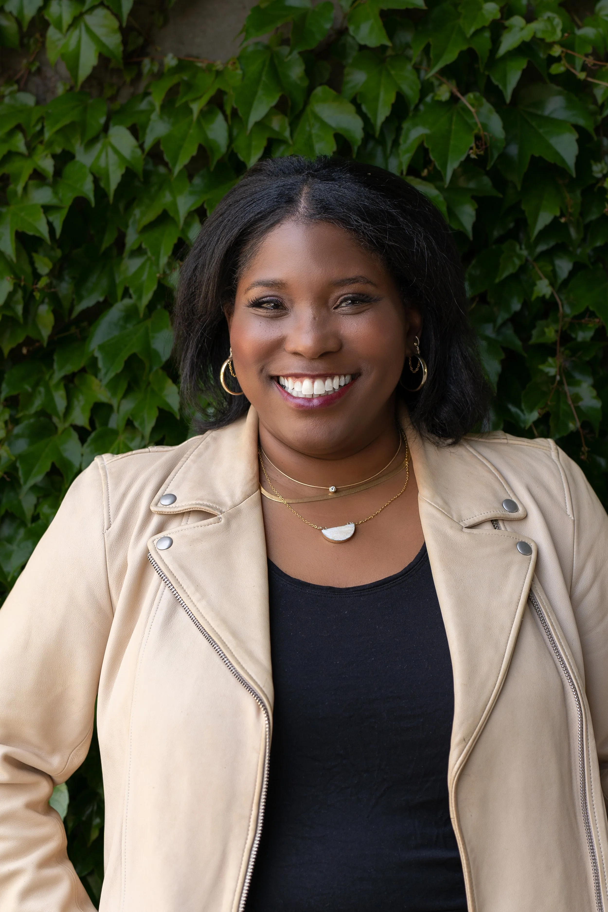 A woman with dark hair, wearing hoop earrings, smiling, dressed in a black top and beige jacket, standing in front of green ivy leaves.