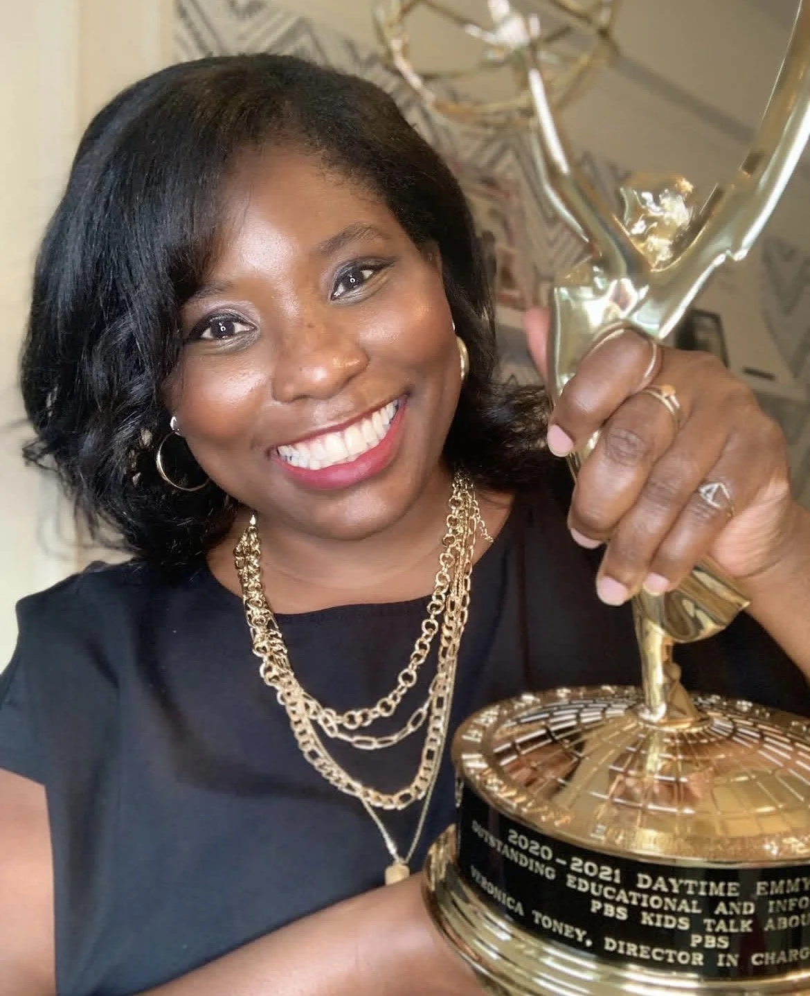 A woman smiling and holding a golden award trophy, wearing a black top with gold jewelry, in a room with patterned wallpaper.