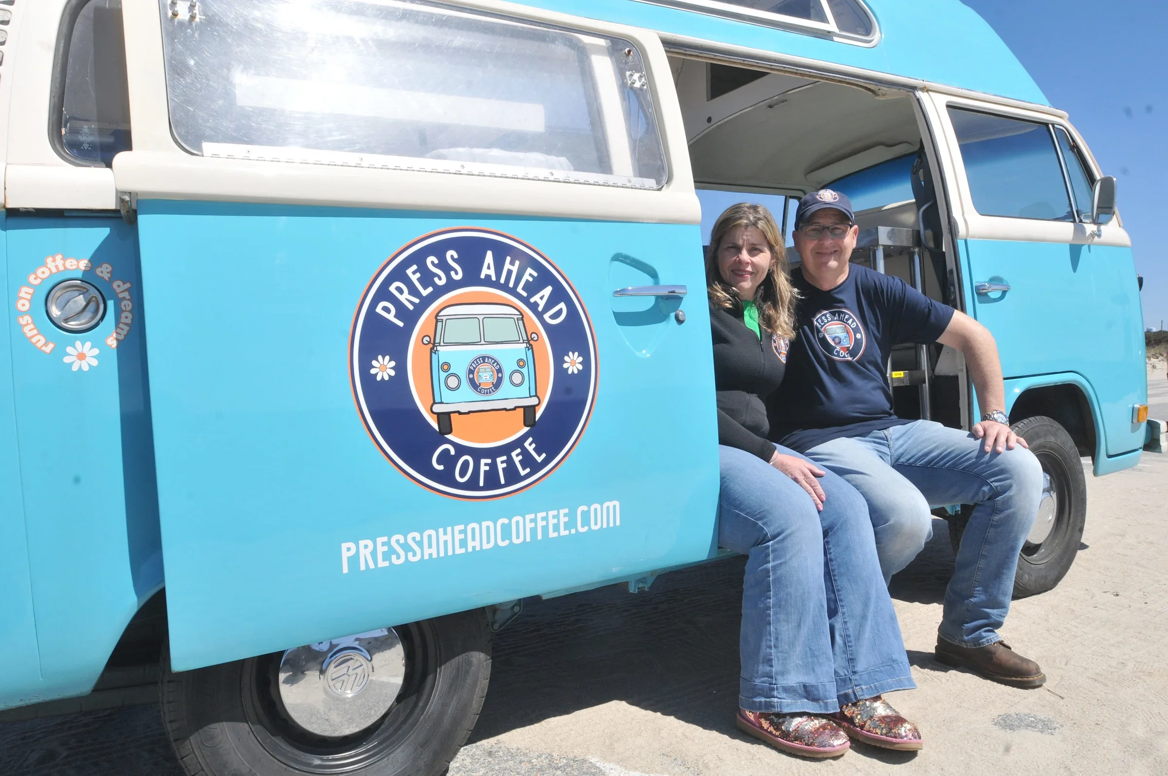 Two people sitting on the edge of a vintage blue food truck with the logo 'Press Ahead Coffee,' which features a stylized blue camper van. They are outdoors on a sunny day, with a clear blue sky in the background. The woman has light brown hair and is wearing a black jacket, while the man is wearing glasses, a cap, and a navy blue shirt. The truck has a website URL pressheadcoffee.com on its side.