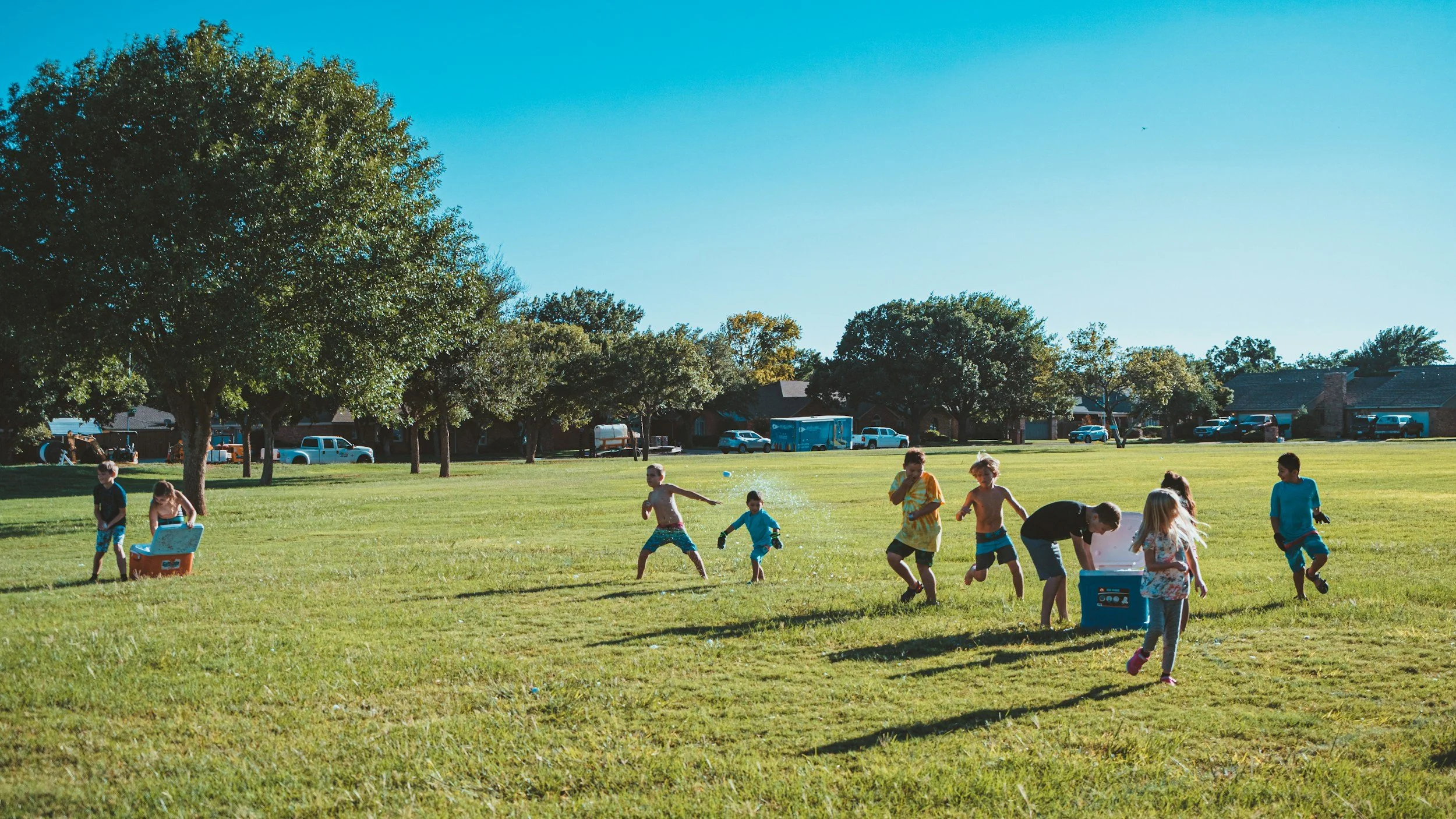 Children playing and running in a grassy park on a sunny day, some near coolers, with trees and houses in the background.