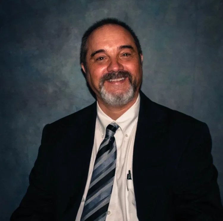 A smiling man with short dark hair, a beard, and a mustache, wearing a black suit, white shirt, and striped tie, sitting in front of a grayish background.