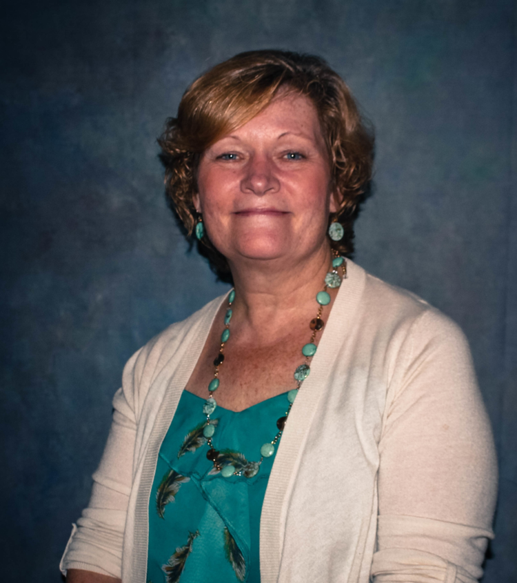 A portrait of a woman with short, curly hair, wearing a turquoise blouse with a feather pattern, a light beige cardigan, and jewelry including turquoise earrings and a matching necklace, against a dark gradient background.