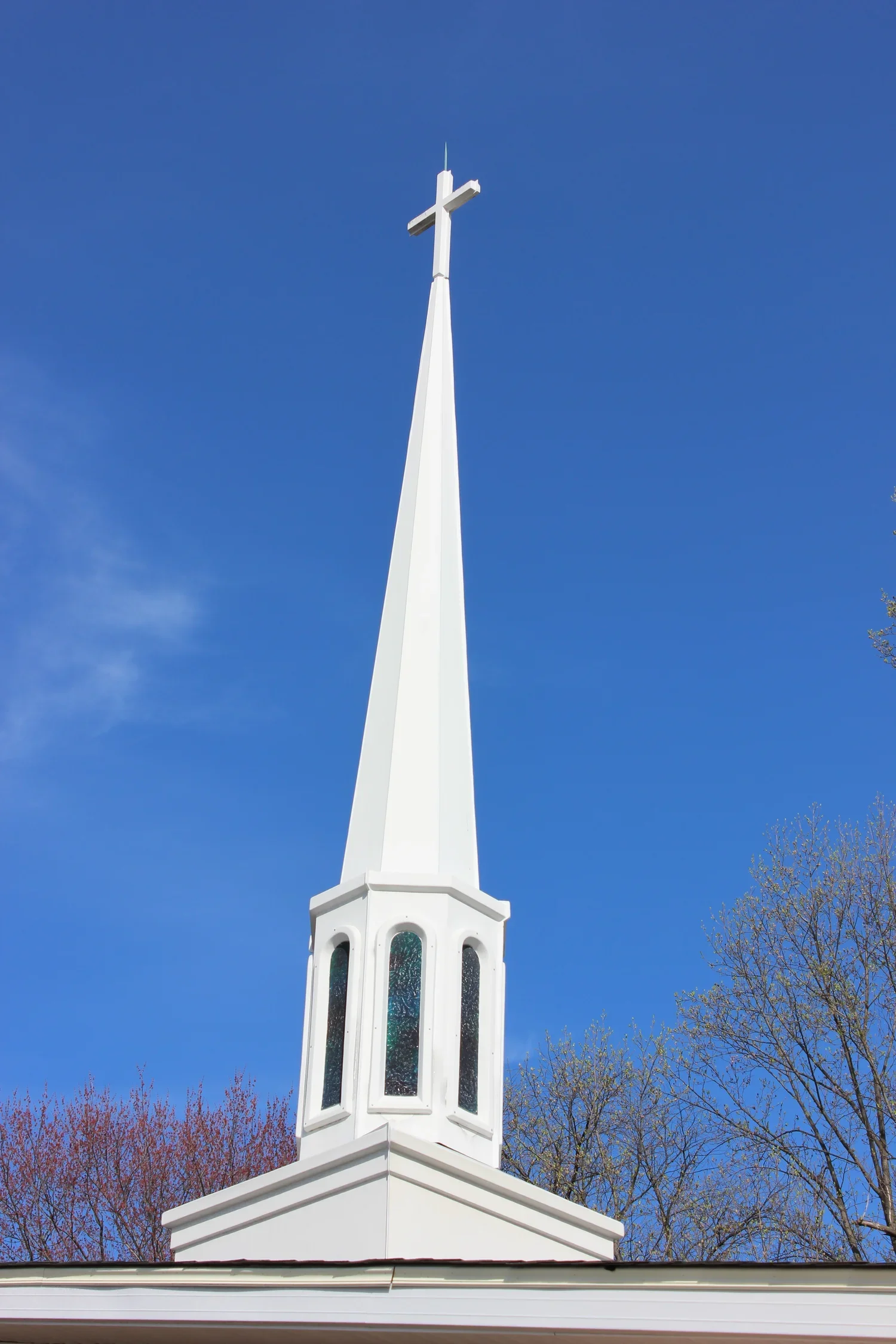 The steeple of a white church building with a cross on top against a clear blue sky, with some trees in the background.