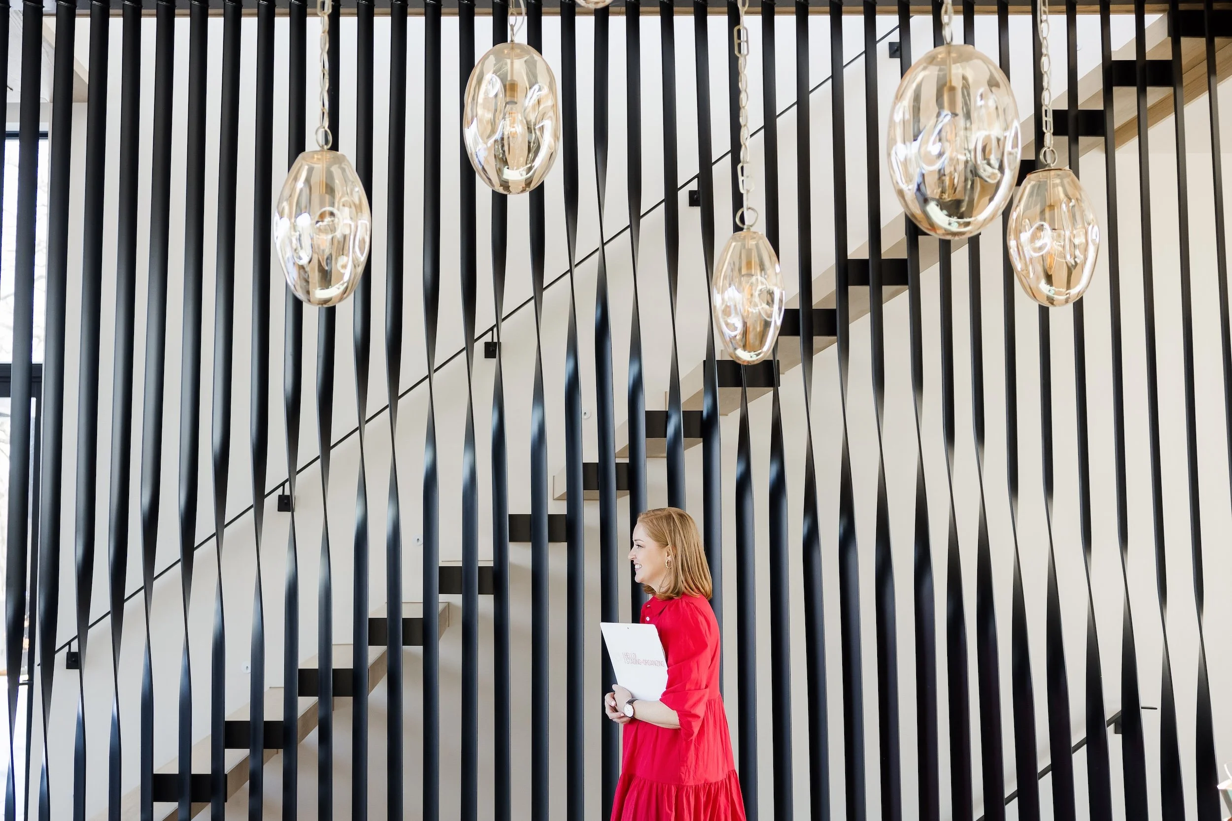 Ashley walking through a foyer area with steps in the background. She is holding a checklist and wearing a red dress.