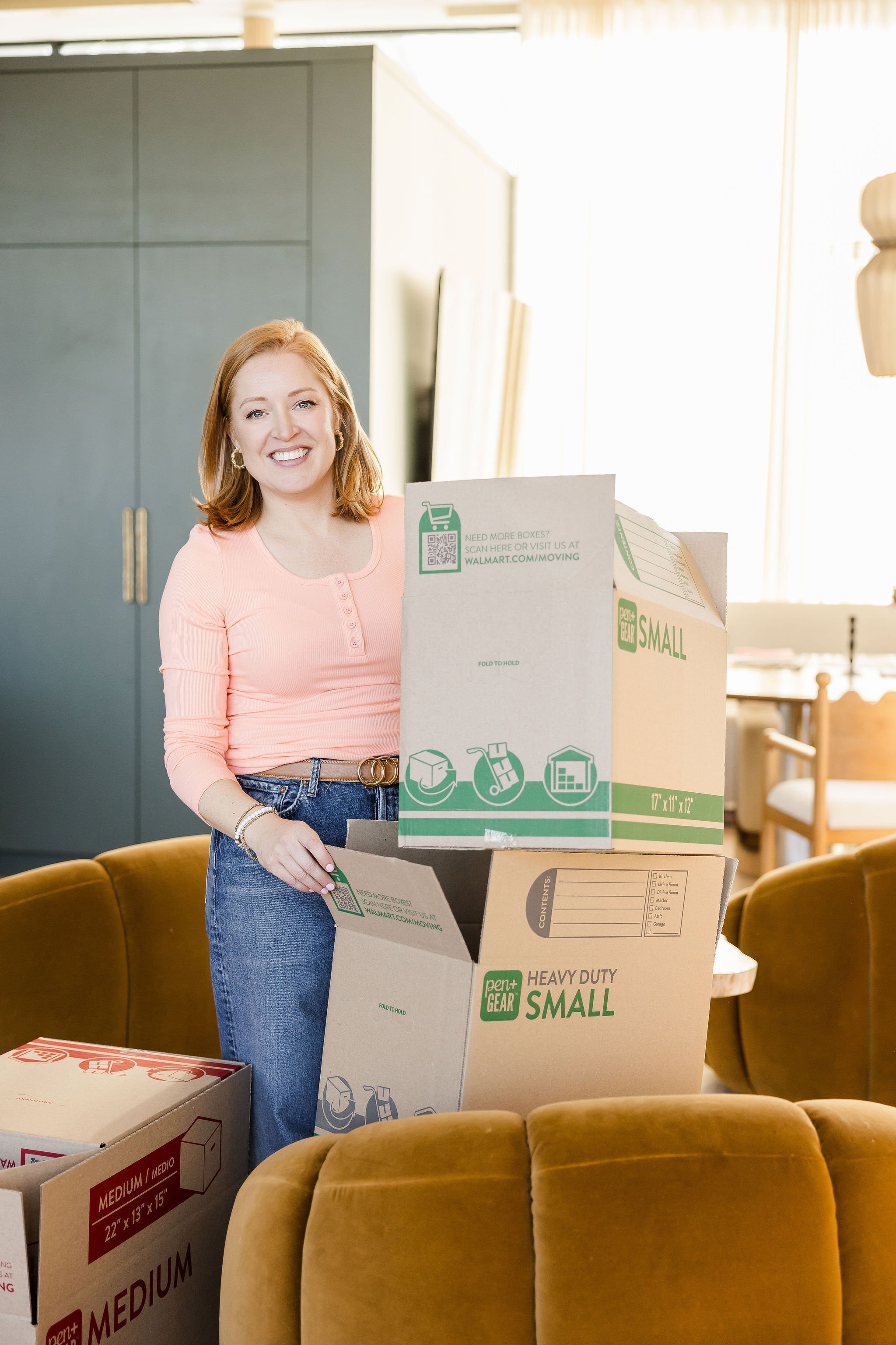 Ashley in a coral shirt and jeans unpacking two boxes and smiling at the camera