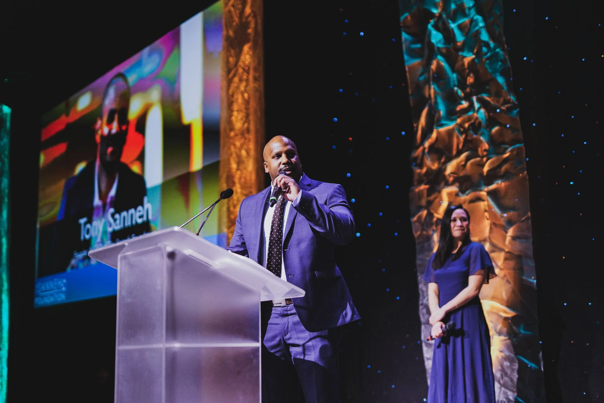 A man in a blue suit speaking at a podium on a stage with a microphone, with a woman in a blue dress standing behind him. There is a large screen displaying his name, Tony Sanneh, and a backdrop with abstract textured panels and star-like lights.