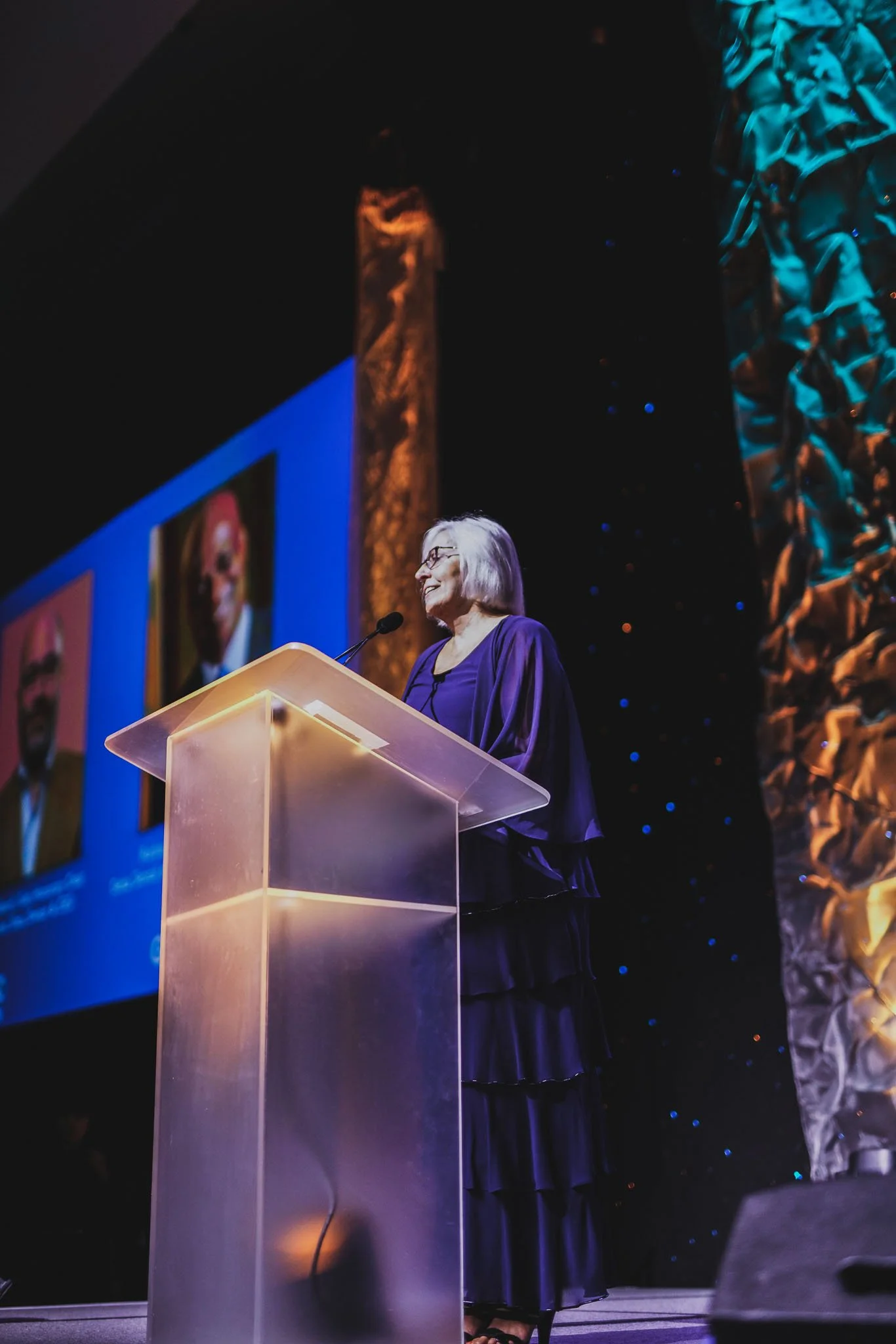 An elderly woman with gray hair and glasses giving a speech at a podium on stage. Behind her, a large screen displays two framed photographs of men. The stage is decorated with gold and blue accents, and she is wearing a flowing purple dress.