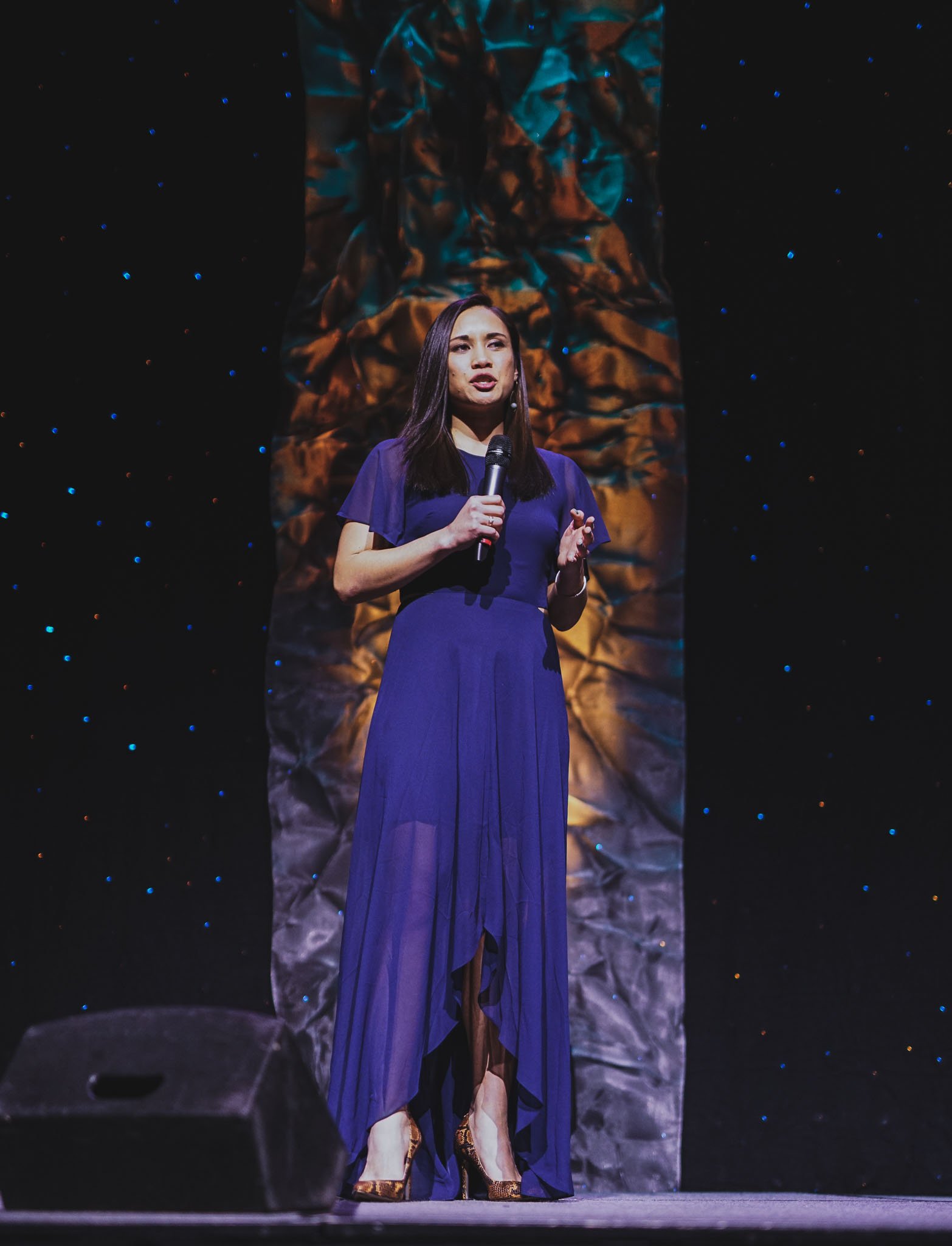 A woman in a blue dress speaking into a microphone on stage with a dark background and textured backdrop.