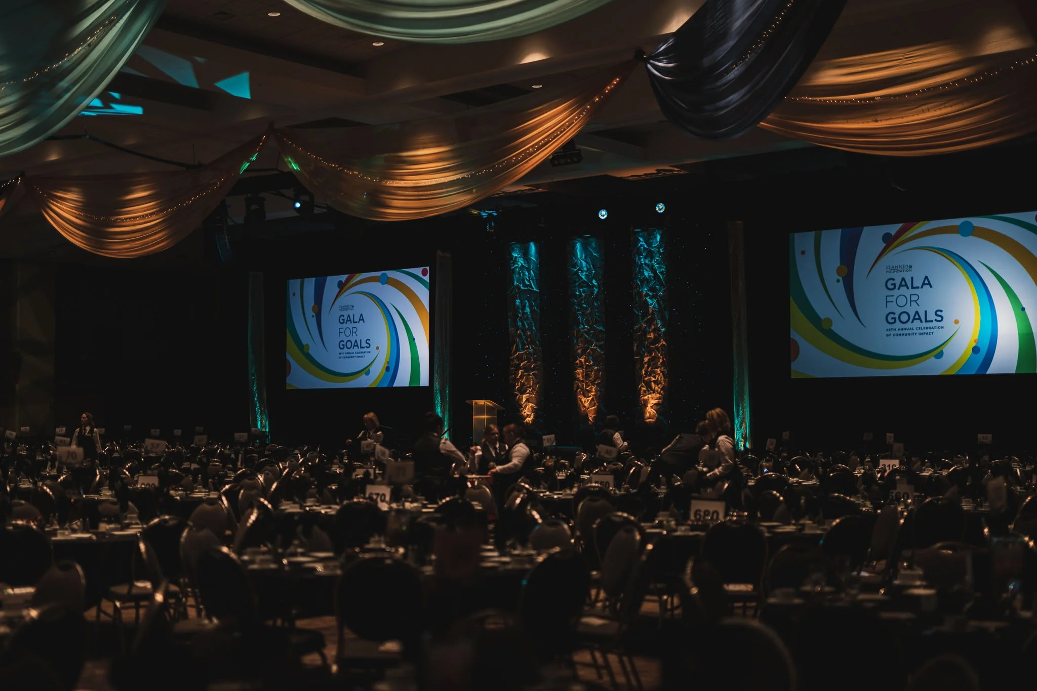 A dimly lit ballroom set up for a gala event with round tables and chairs, large screens displaying 'Gala for Goals,' and decorative draping on the ceiling.