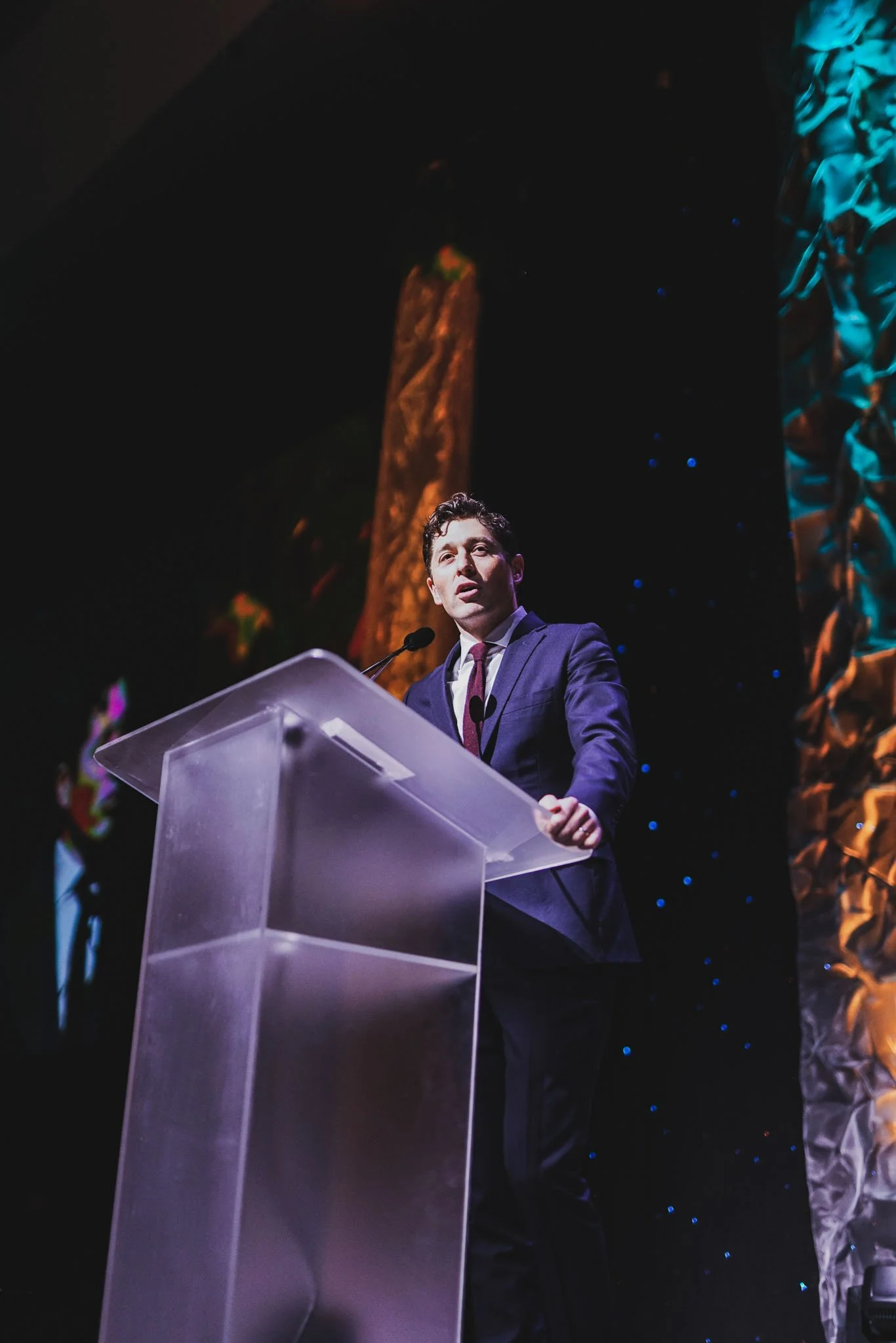 A man in a dark suit and tie giving a speech at a podium on a stage with colorful decorative backdrops.