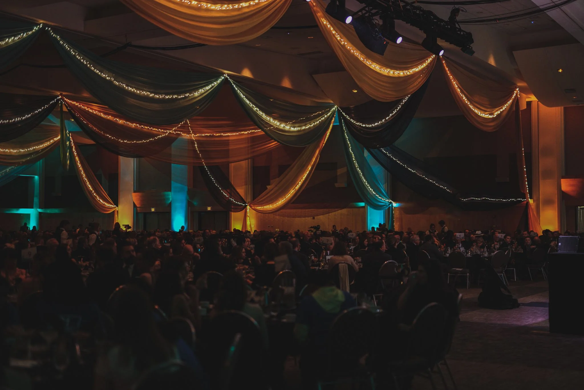 A large banquet hall with draped fabric and string lights hanging from the ceiling, filled with round tables and attendees.