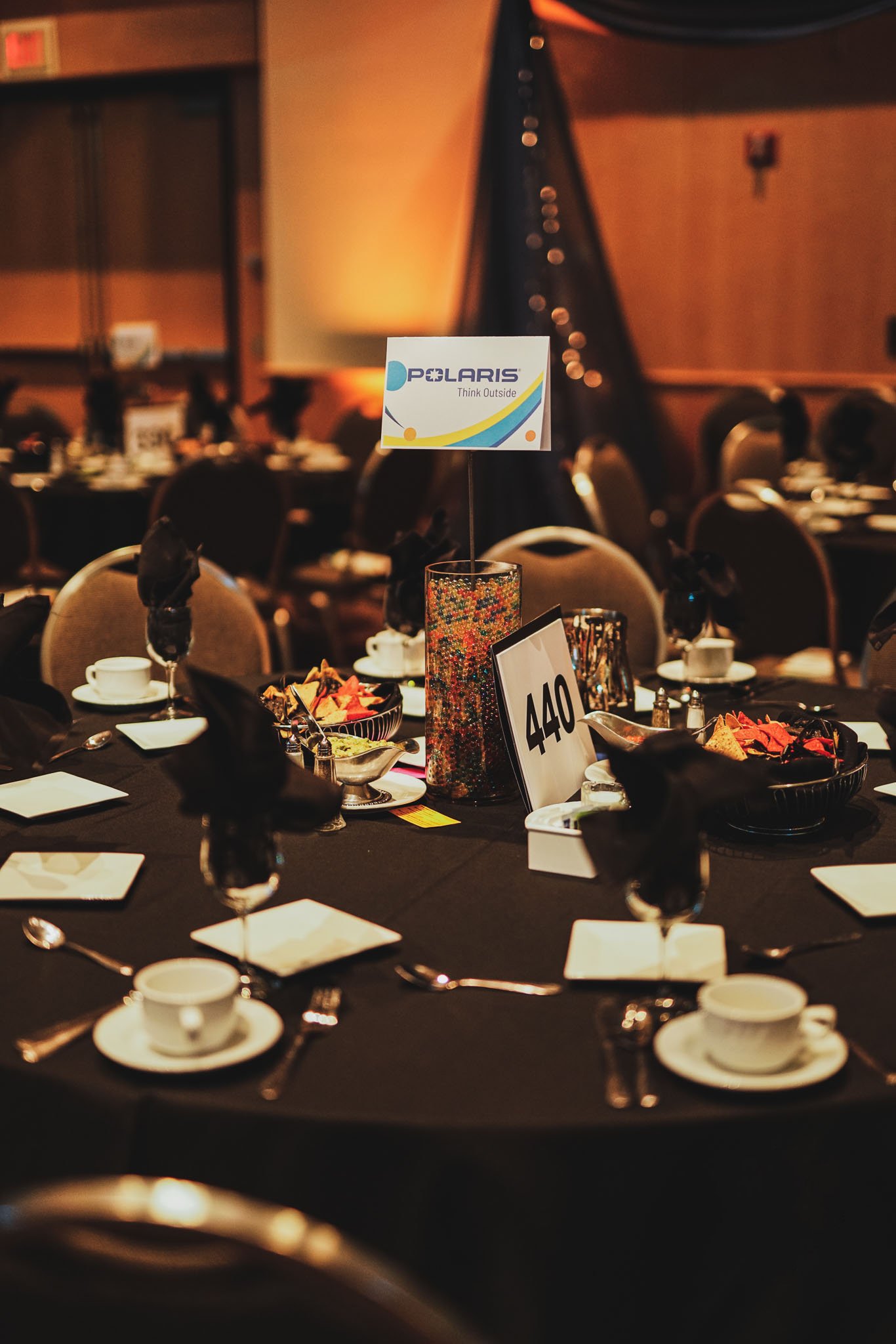 A round banquet table set with black napkins, white cups and saucers, small plates, and utensils. In the center, there is a colorful beaded centerpiece with a table number '440' and an umbrella with a Polaris sign. The table is located in a dimly lit
