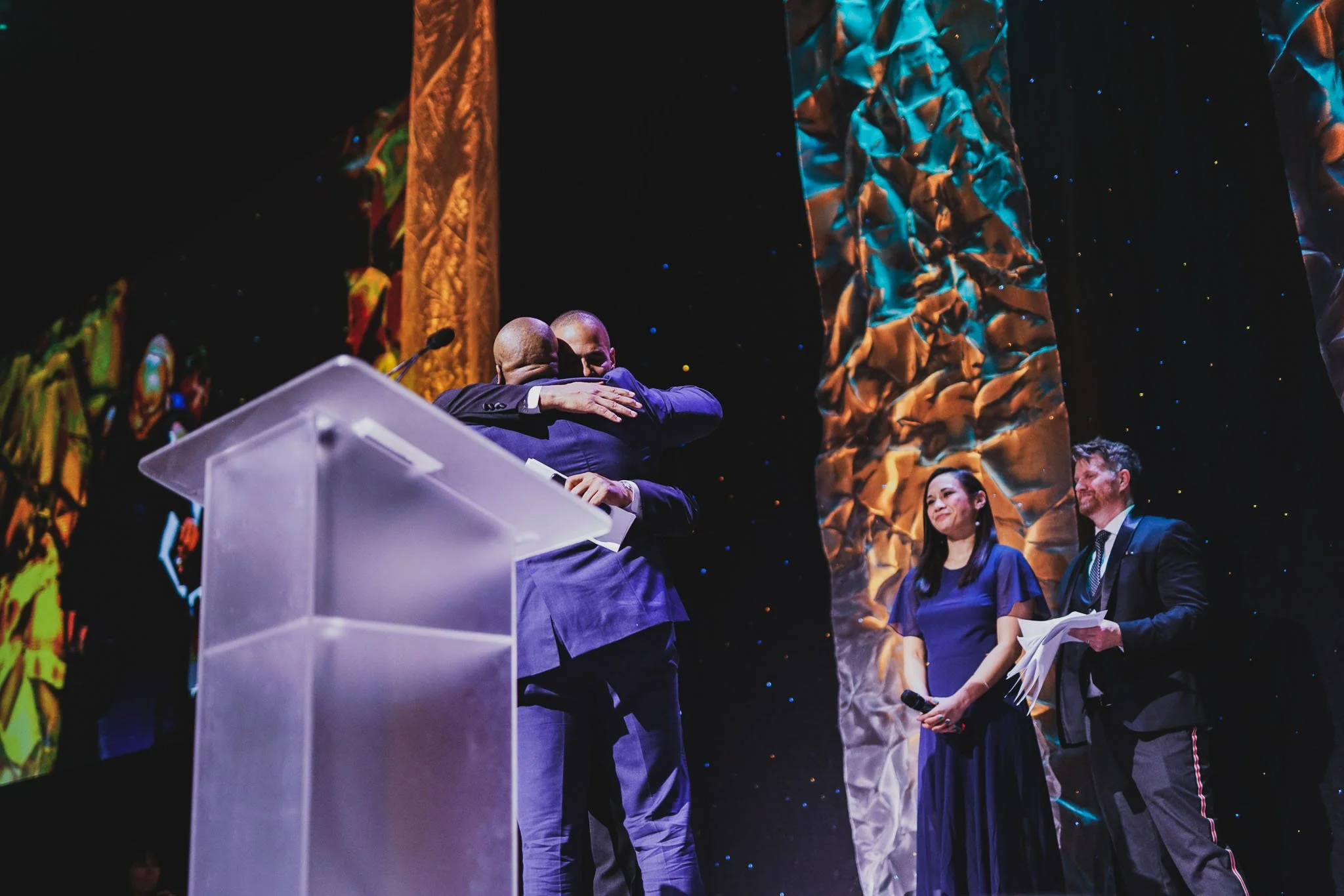 Two men in suits are hugging on stage at an award ceremony, with a woman and a man holding papers standing nearby, against a colorful, textured backdrop.