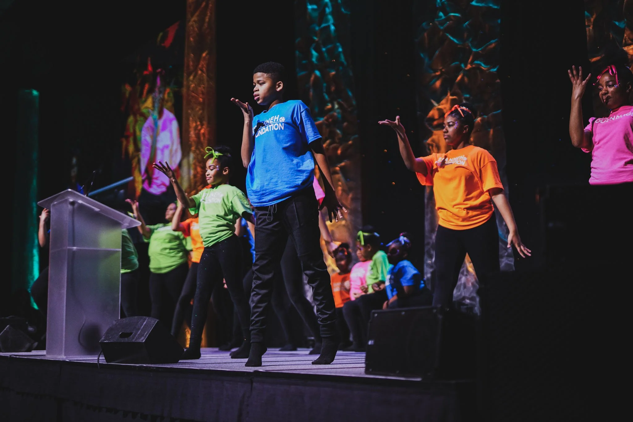 Children performing a dance on stage, wearing colorful shirts in green, blue, orange, and pink, with a decorative background.