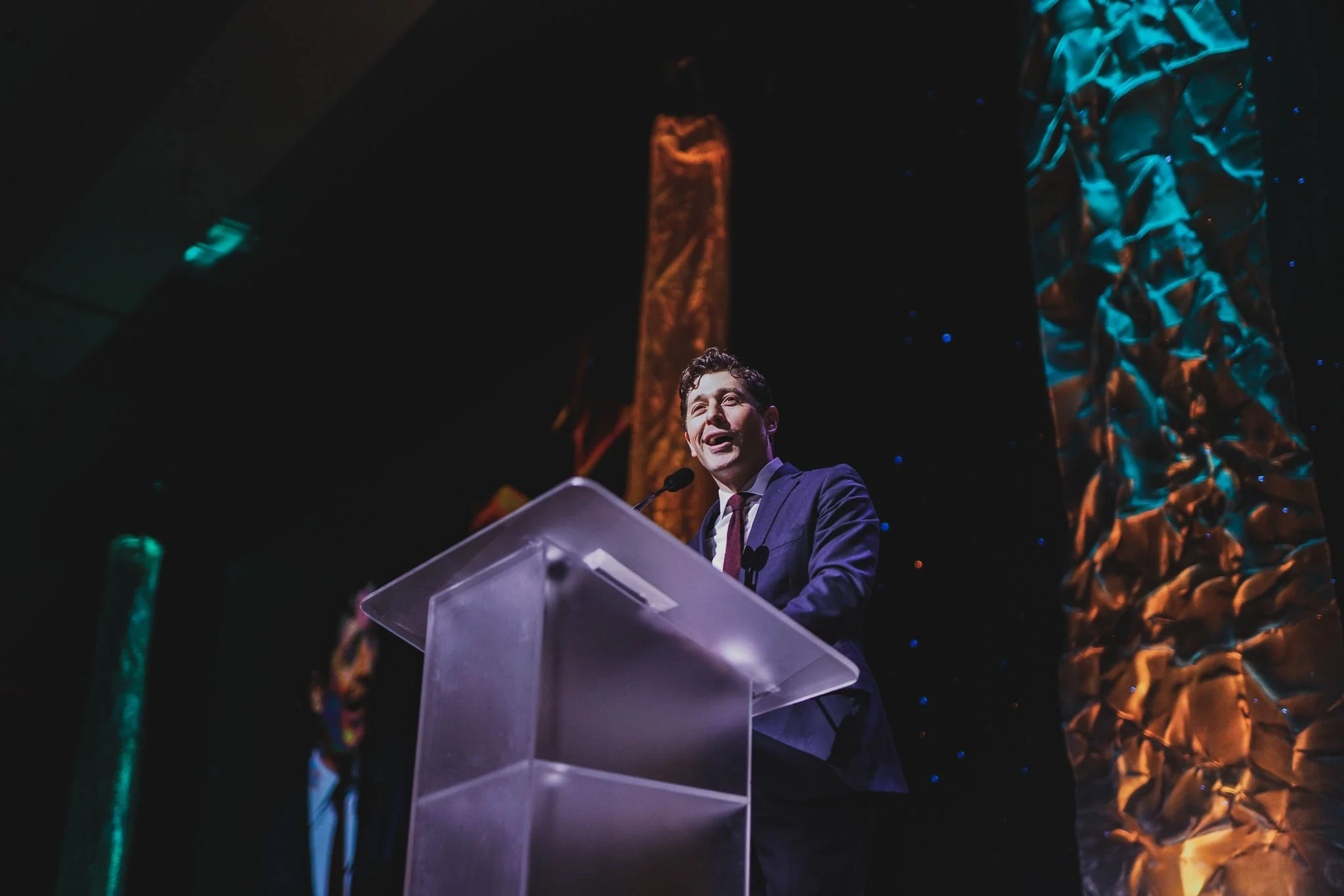 A man in a suit speaking at a podium during an event, with decorative gold and blue fabric drapes behind him.