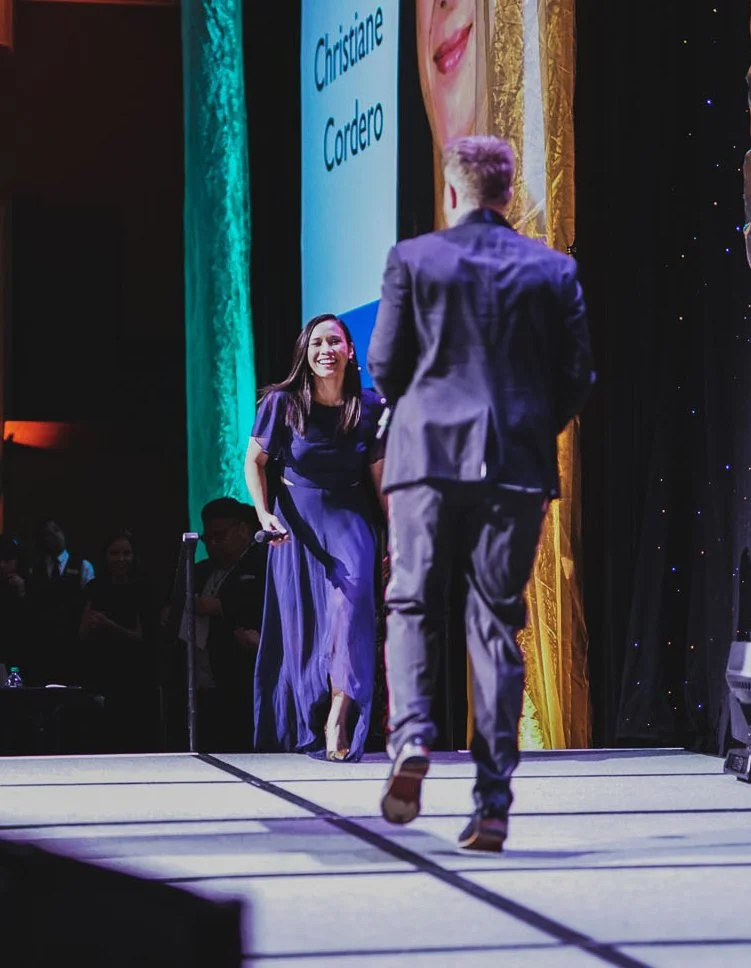 A woman in a purple dress is smiling as she approaches a man in a suit on a stage. The stage has curtains and a large screen displaying the name Cristiane Cardero and her photo.