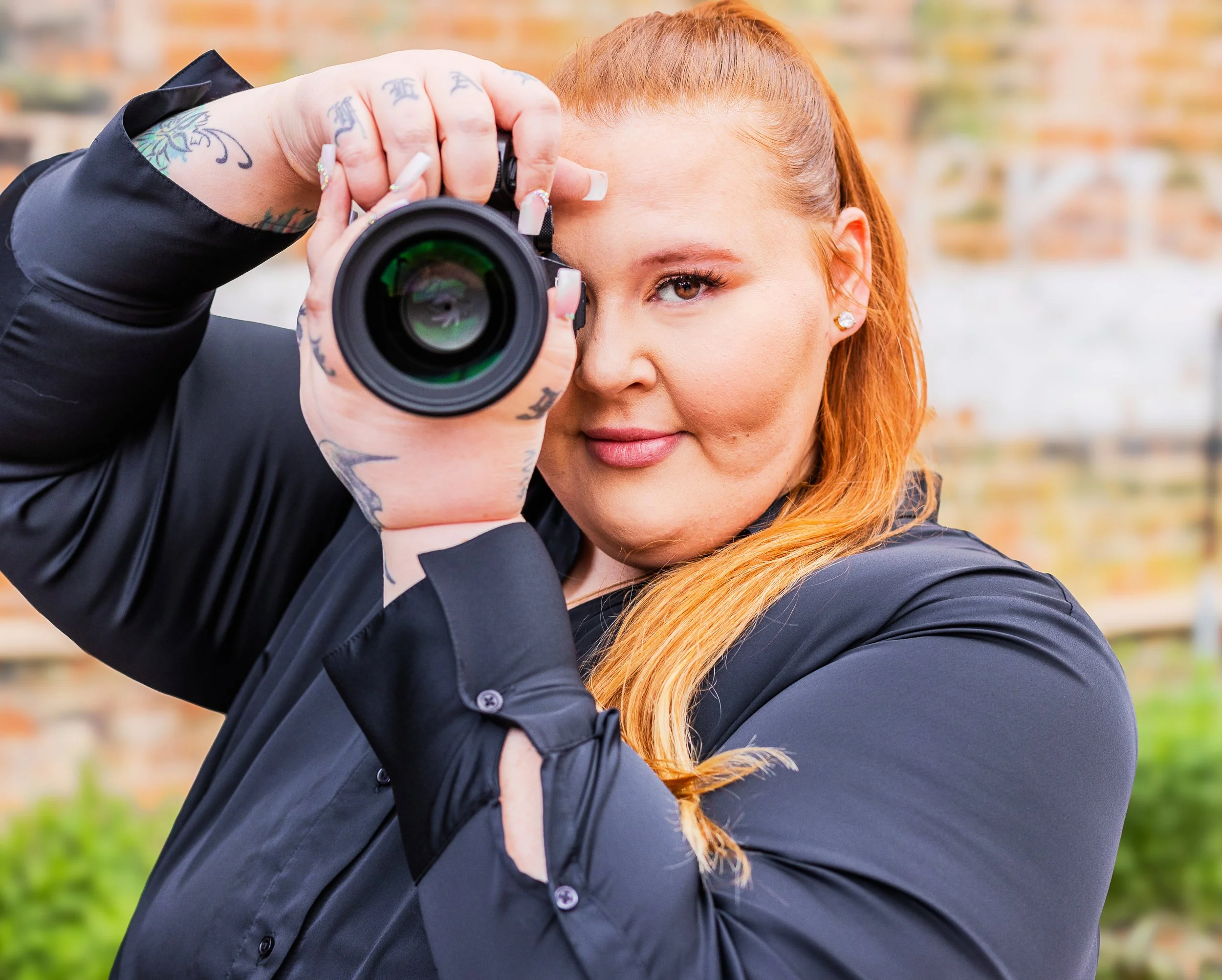 Woman with red hair taking a photo with a camera outdoors, wearing a black jacket, with a brick wall and green grass in the background.