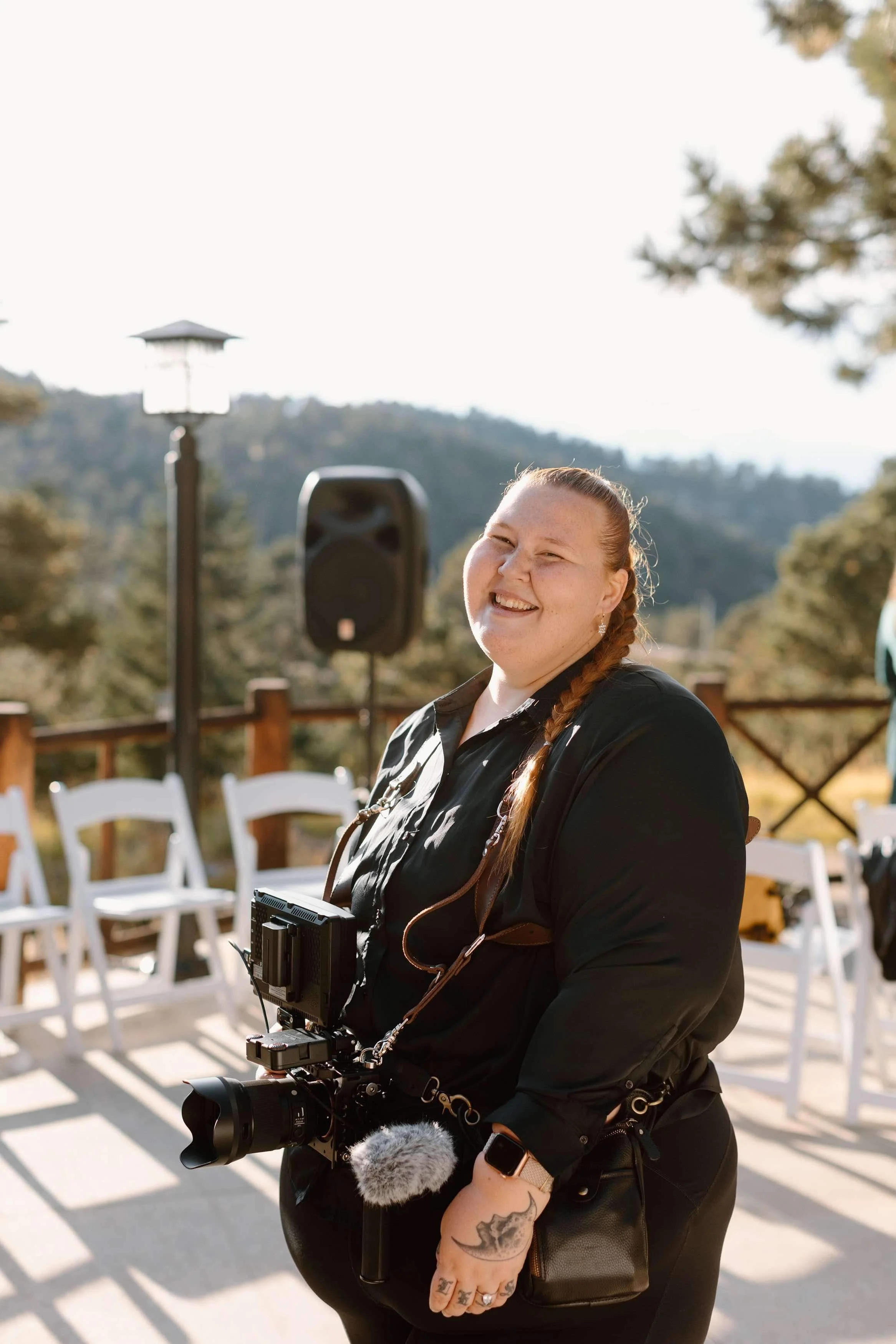 A smiling woman with long braided hair wearing a black jacket, holding a professional camera with a microphone, outdoors in a sunny area with trees and white chairs in the background.