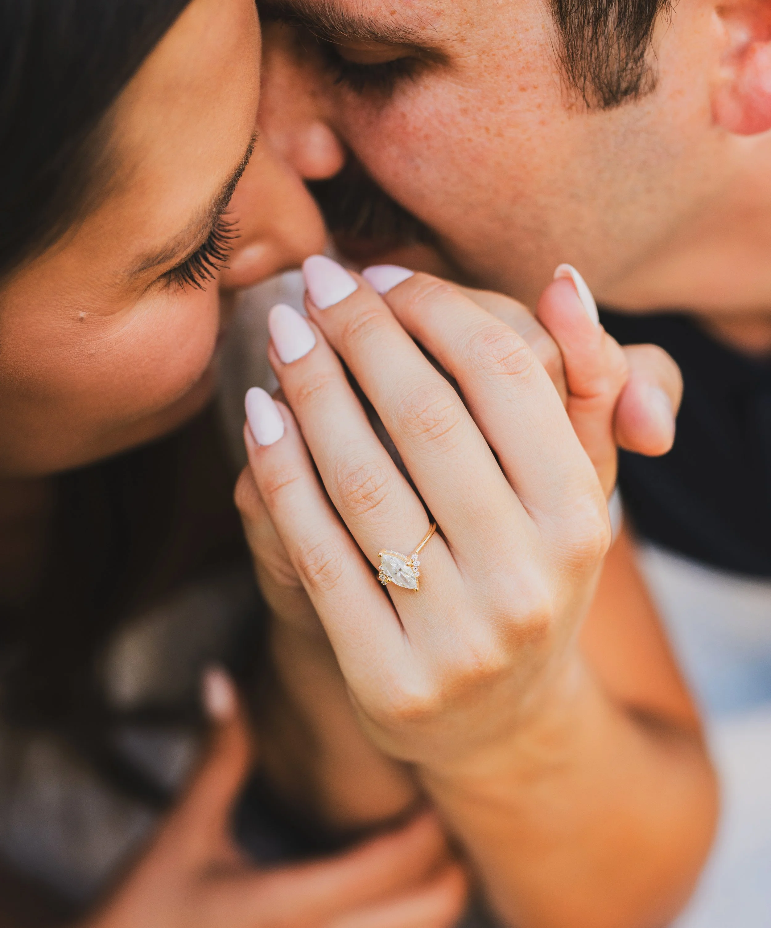 Close-up of a woman with long dark hair and manicured nails kissing a man with closed eyes, showing an engagement ring with a large oval diamond on her finger.