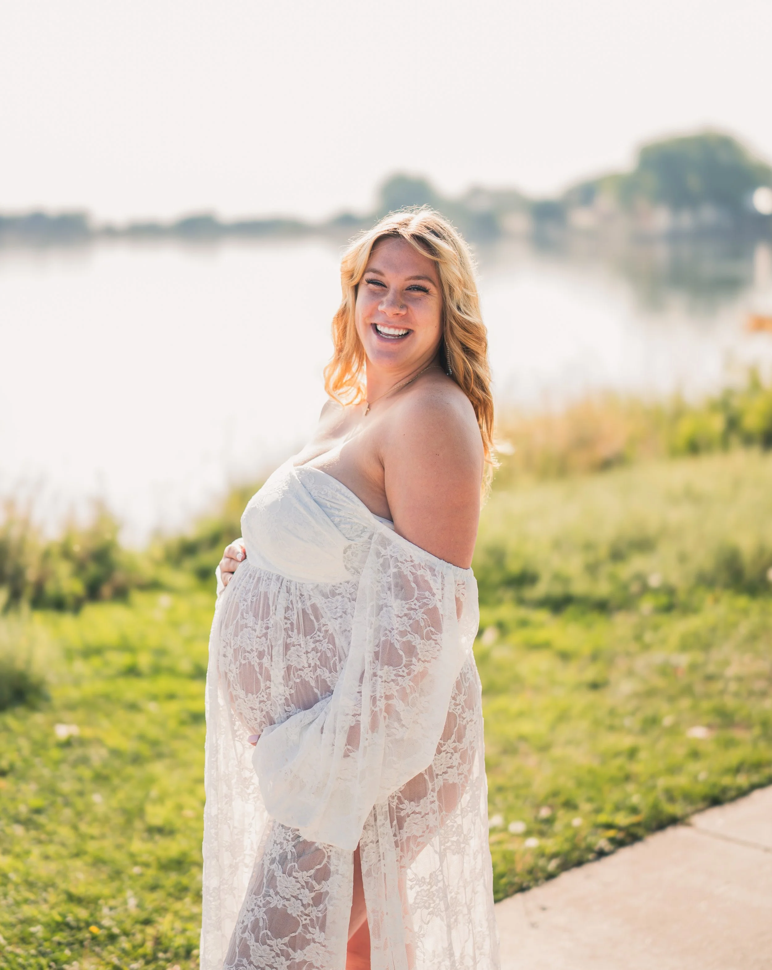 A pregnant woman with blonde hair wearing a white lace dress, standing outdoors near a body of water, smiling and looking at the camera.