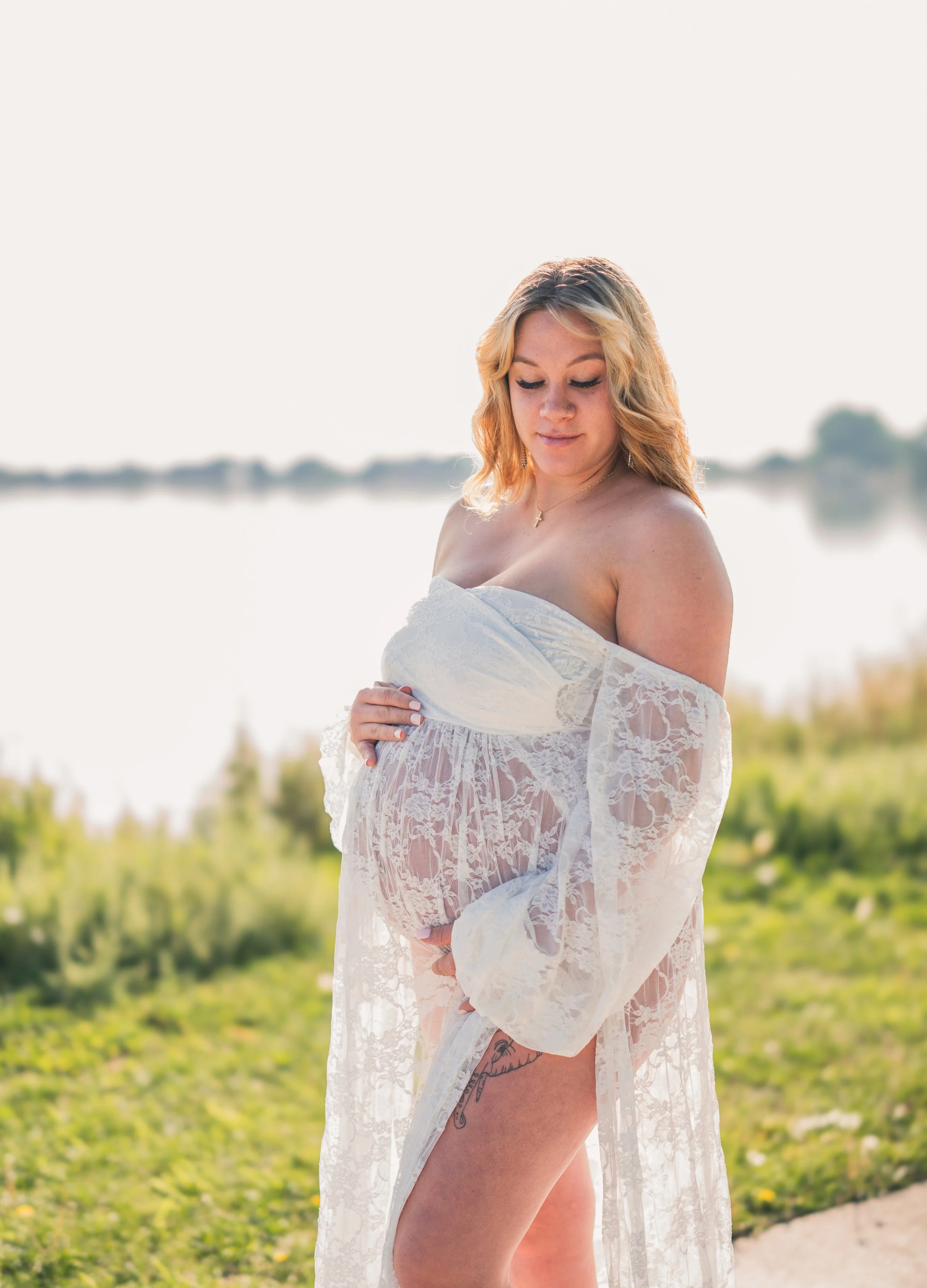A pregnant woman in a sheer lace white dress looking down and standing outside near water with greenery in the background.