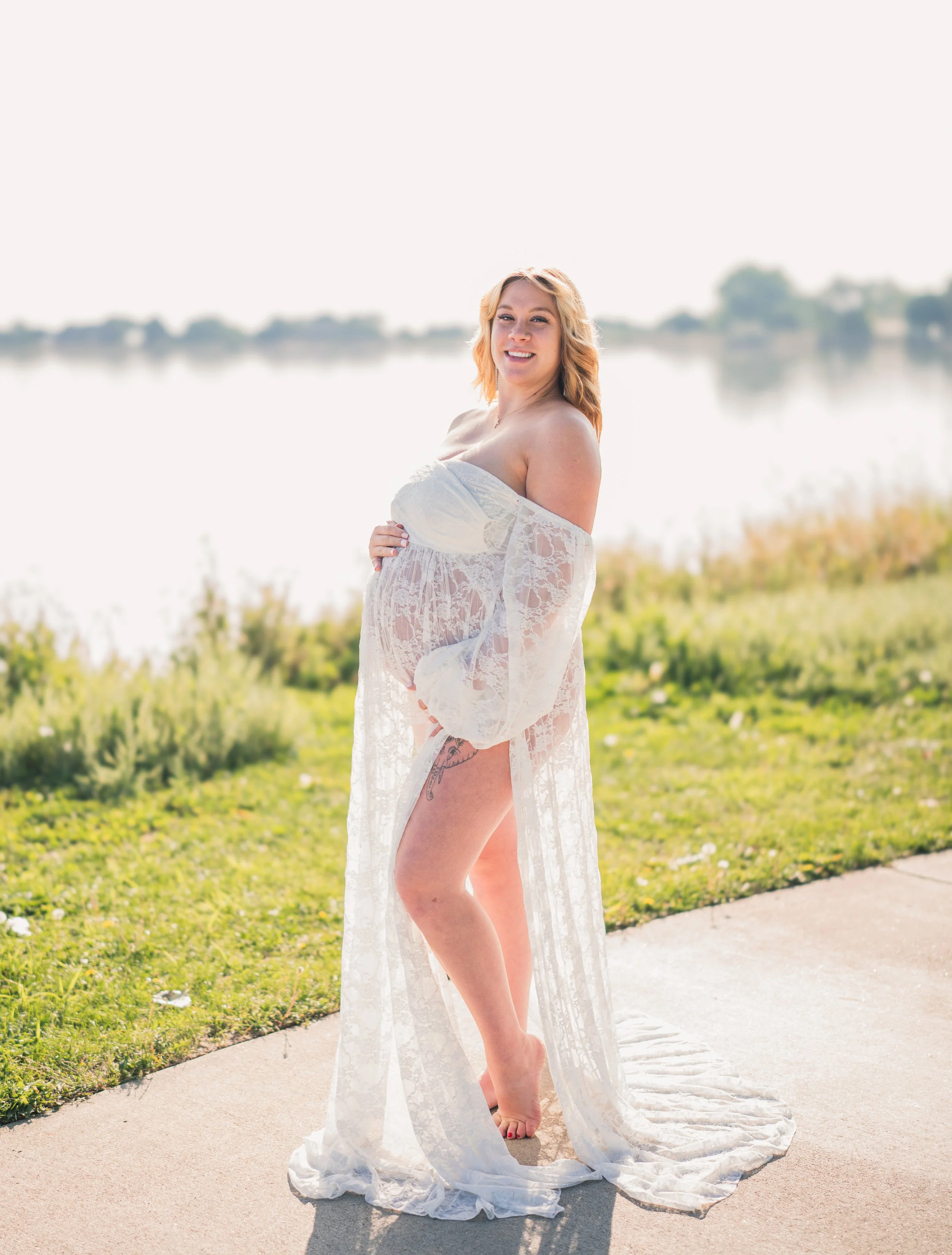 Pregnant woman in white lace dress standing outdoors by a lake, smiling, with greenery and water in the background