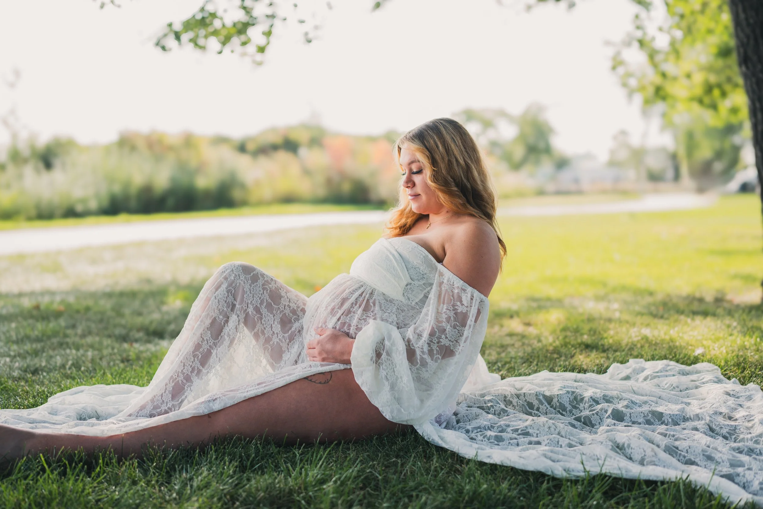 Pregnant woman wearing a white lace dress sitting on the grass under a tree in a park, looking down and holding her belly.