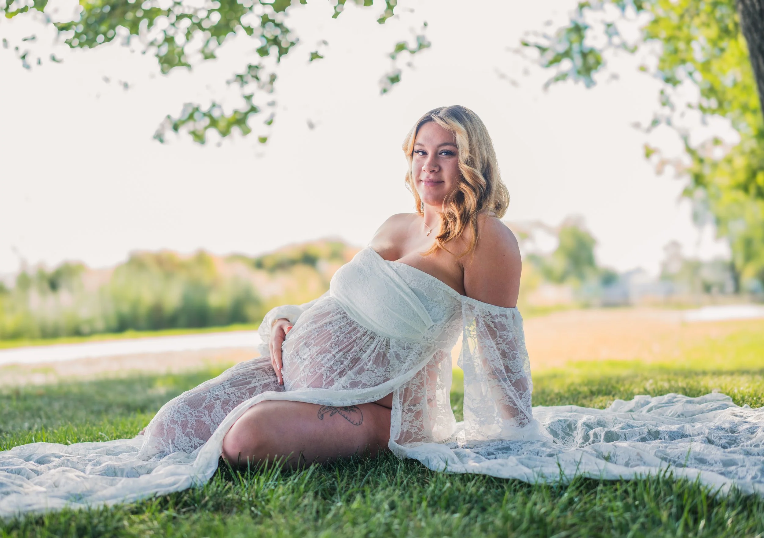 A woman in a sheer white lace dress, sitting on a blanket outdoors in a park, with trees and a lake in the background.