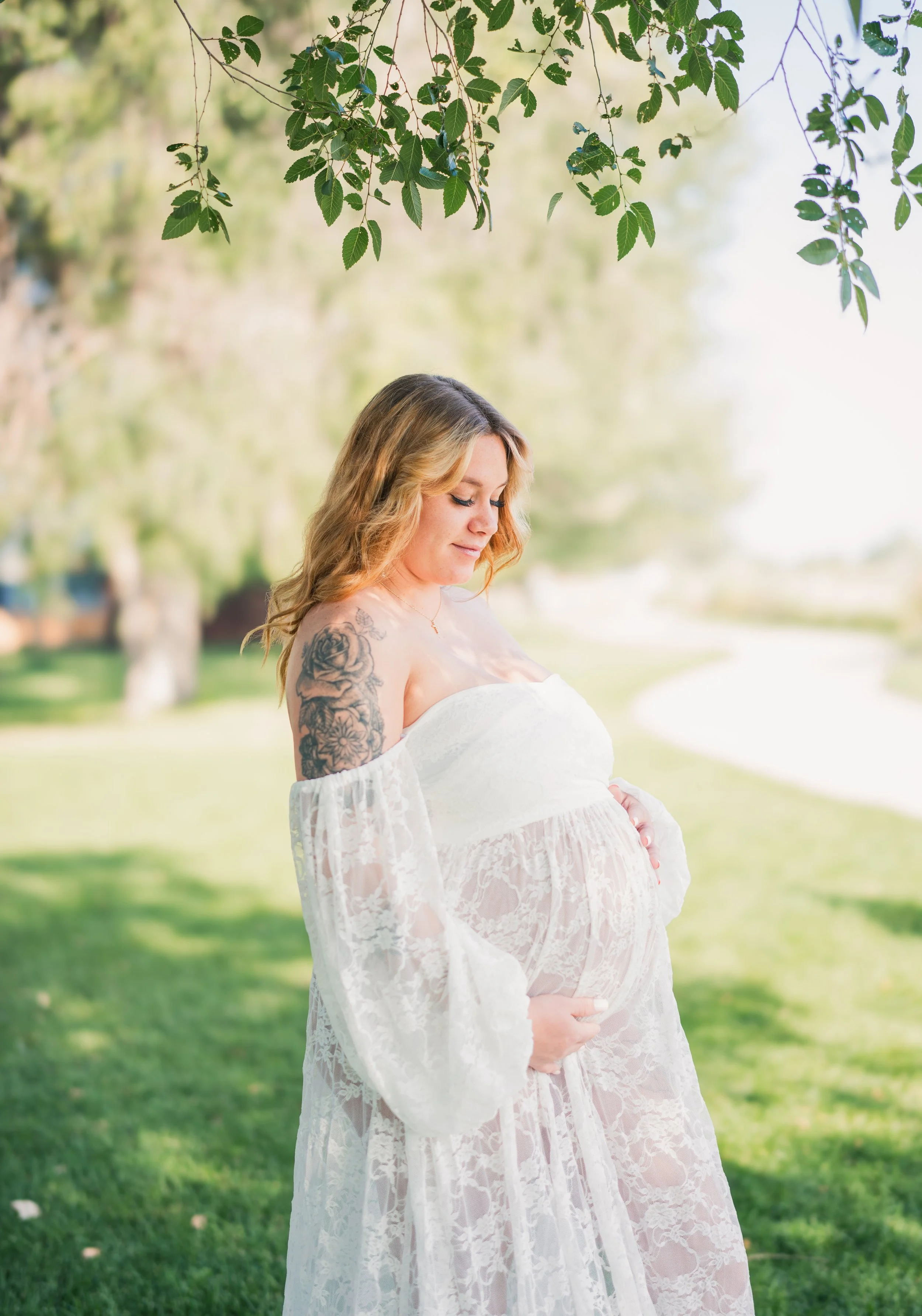 Pregnant woman in a white lace dress holding her belly outdoors under a tree.